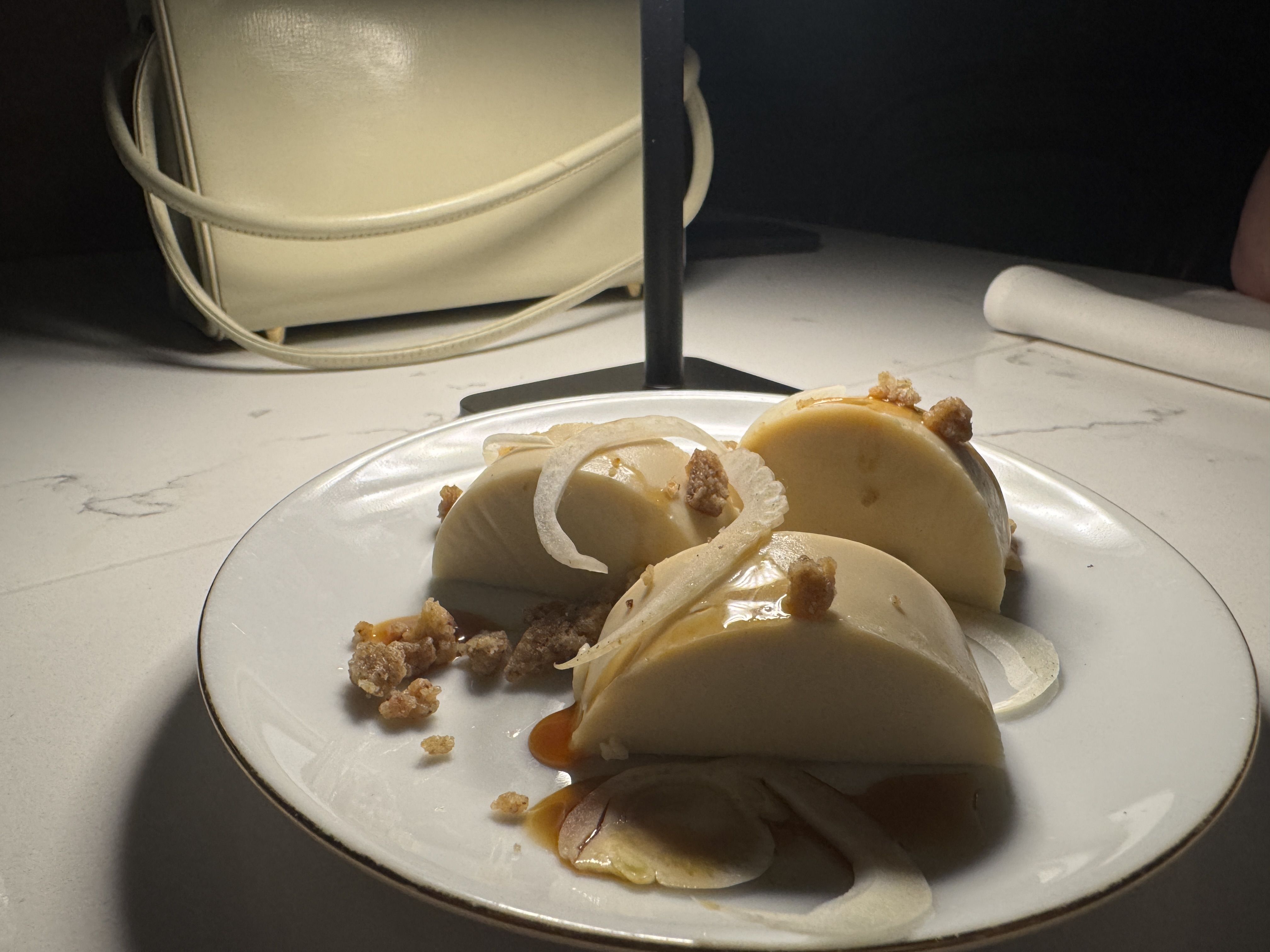 Three smooth, cream-colored dessert halves on a white plate with caramel sauce, thinly sliced onions, and small crunchy bits, set against a marble table and a cream handbag in the background.