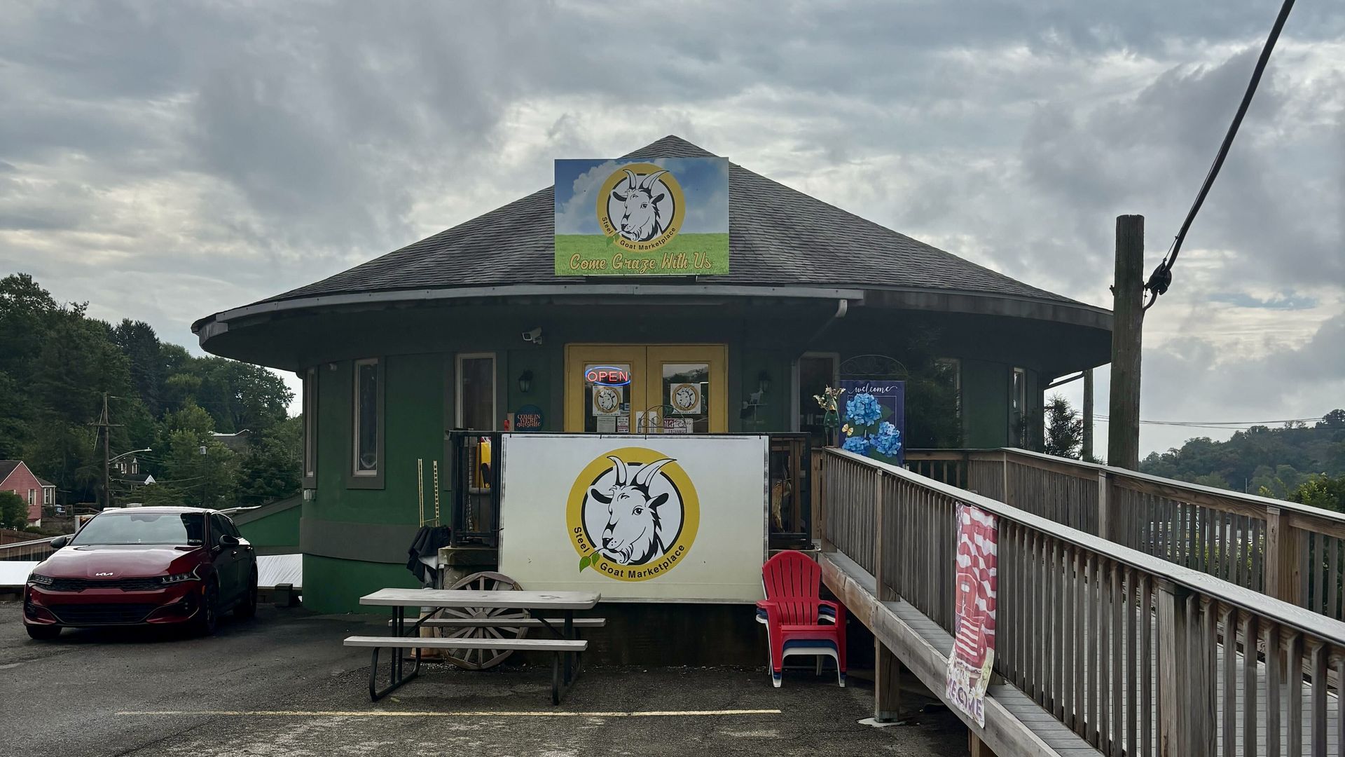 Green round building with gray roof labeled Steel Goat Marketplace, yellow doors, picnic table, red chairs, and a red car parked to the left under a cloudy sky.