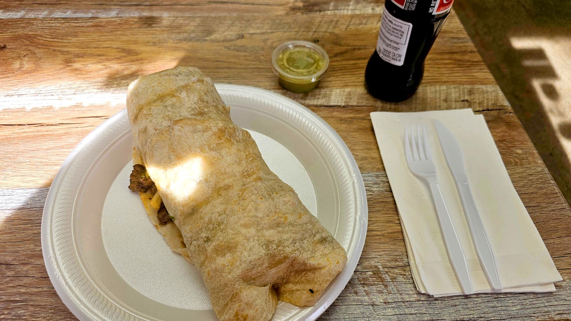 Burrito on a white disposable plate, small container of green salsa, plastic fork and knife on napkins, and a dark soda bottle on a wooden table.