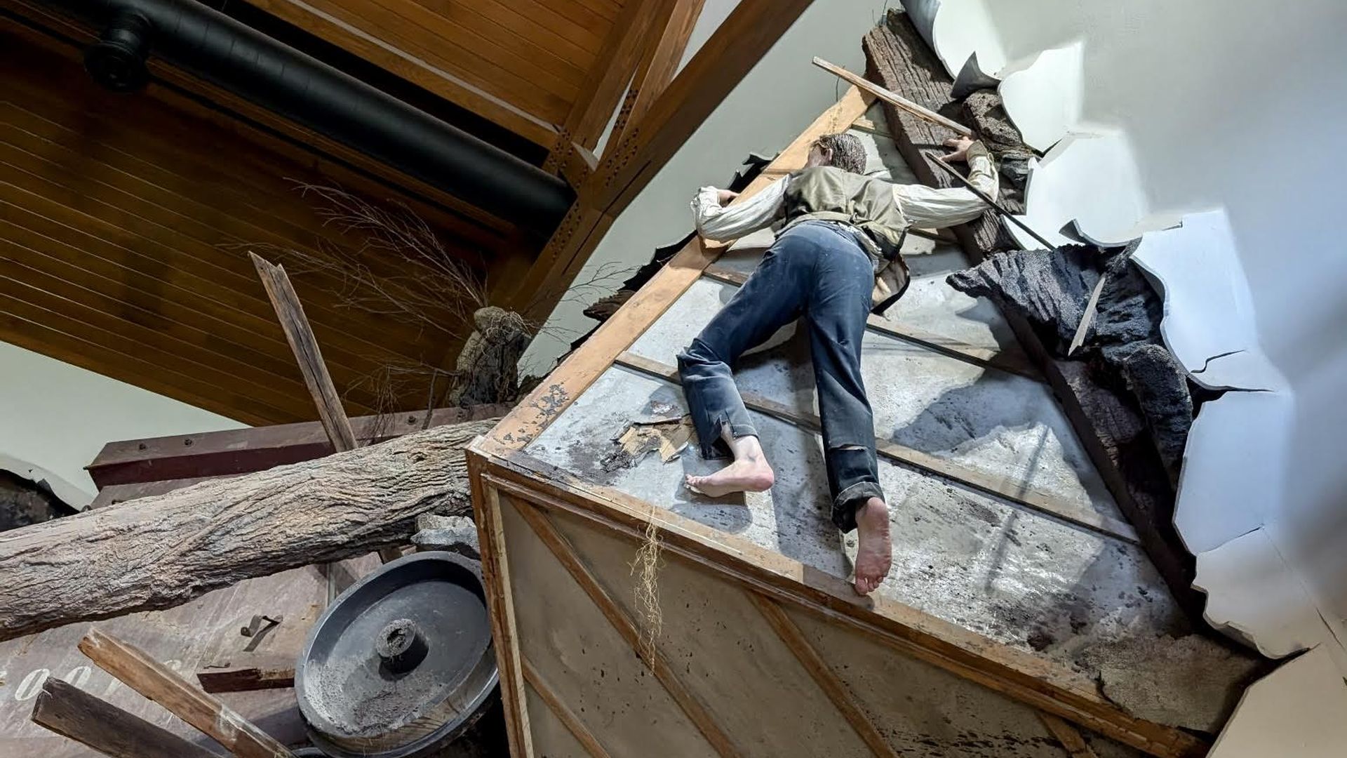 A figure dressed in vintage clothes lies face down on a slanted concrete surface with scattered debris and a large tree trunk nearby in an indoor setting with wooden beams and white walls.