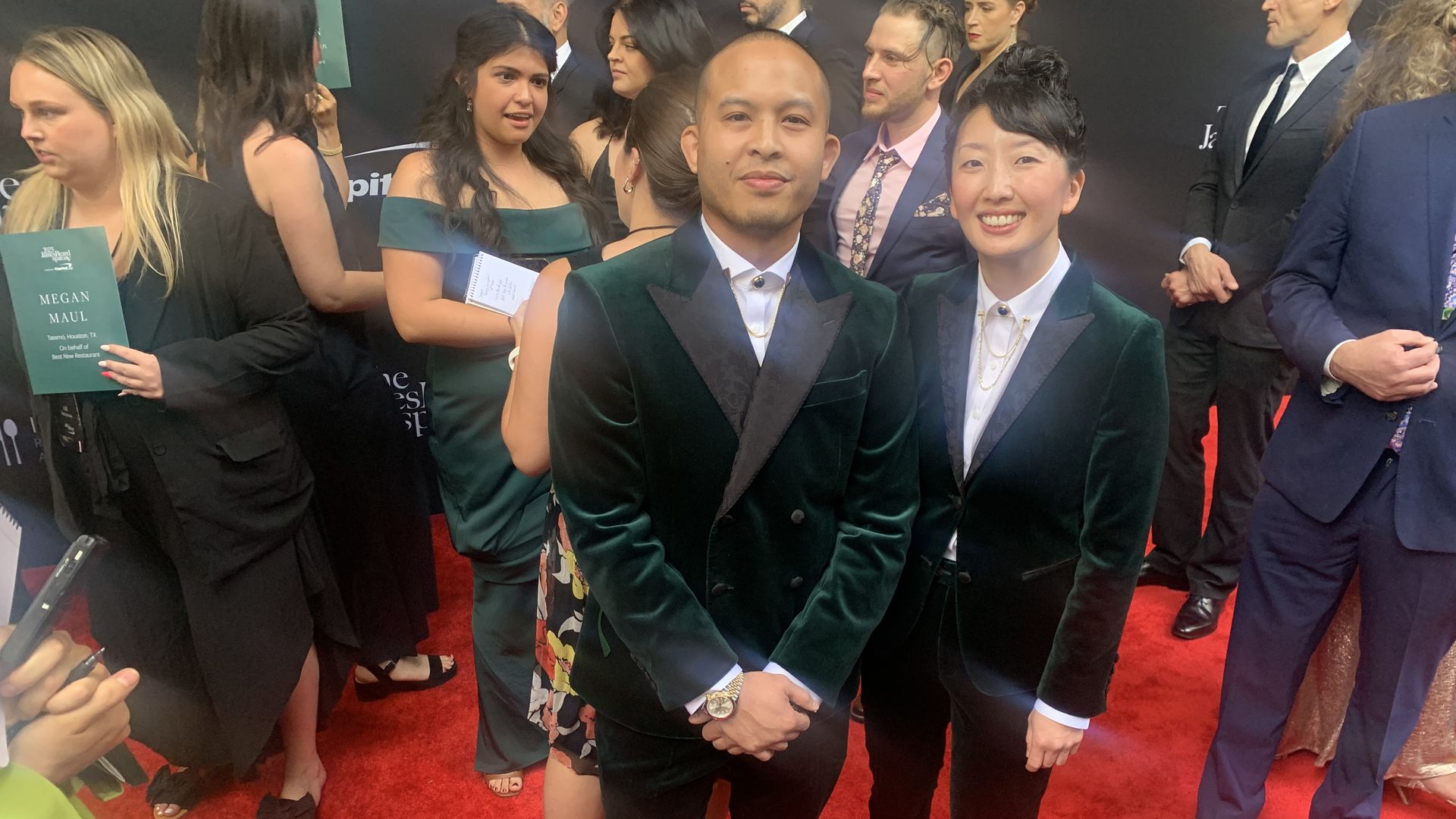 Photo of two people in velvet tuxedos pose on a red carpet. 