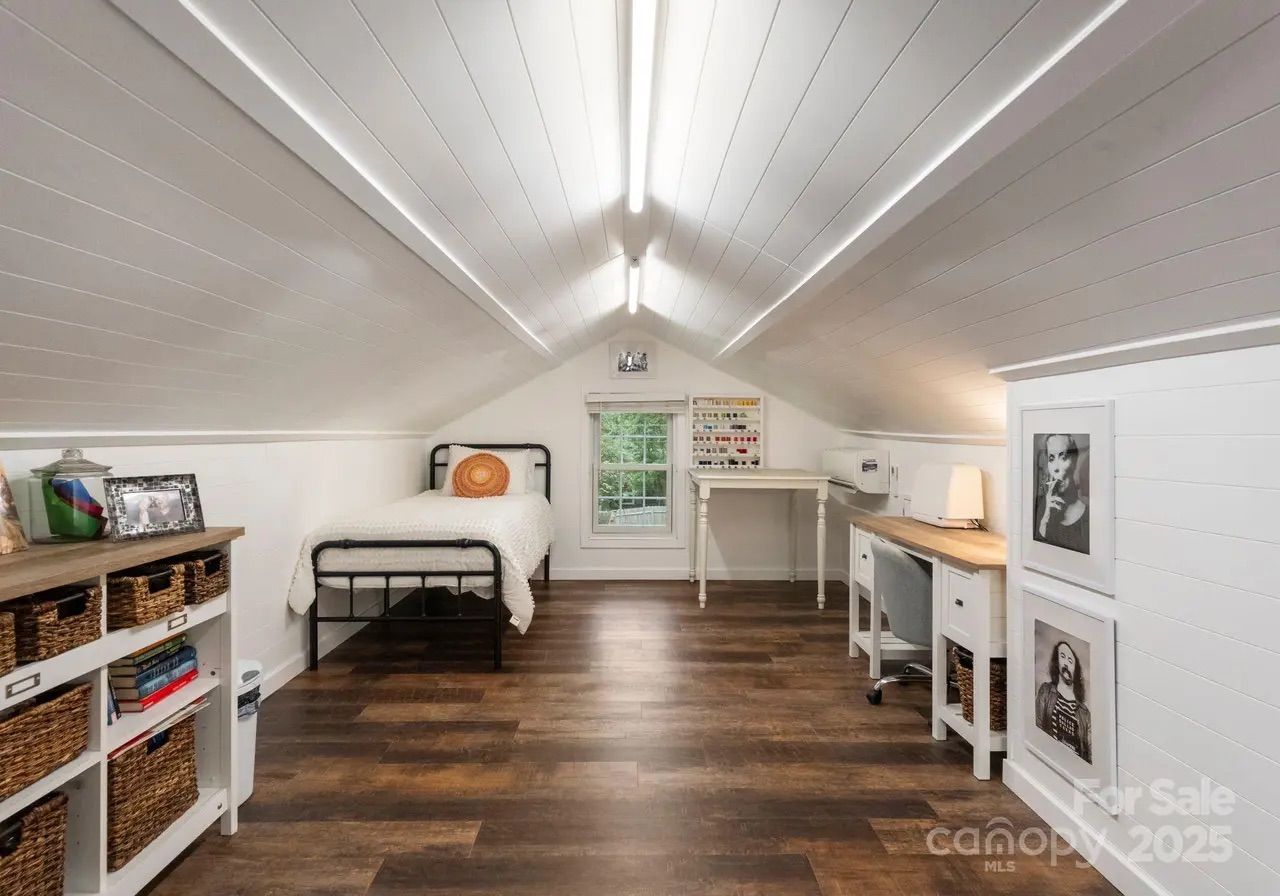 Bright attic room with white shiplap walls and ceiling, dark wood floor, a single bed with orange round pillow, workspace with sewing machines, and woven baskets on shelves.