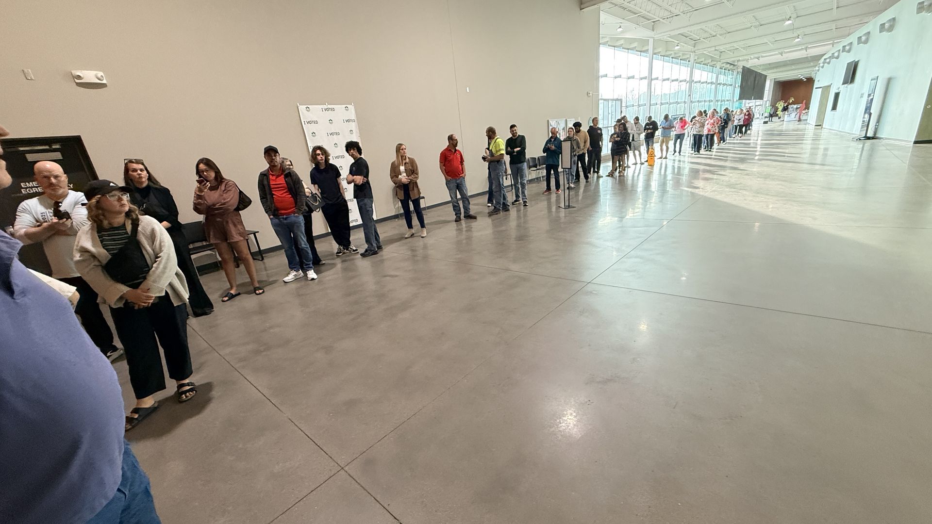 A long queue of diverse people waiting to vote along a beige wall in a bright, modern atrium with glass walls to the right; a banner reads "I VOTED" near the front.