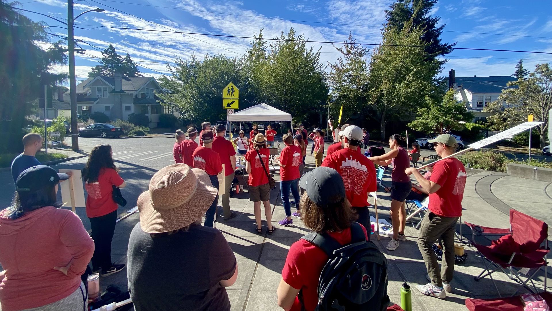 Group of people in red shirts gather around a white tent on a sunny street, with a yellow pedestrian sign, power lines, and tall green trees against a bright blue sky.