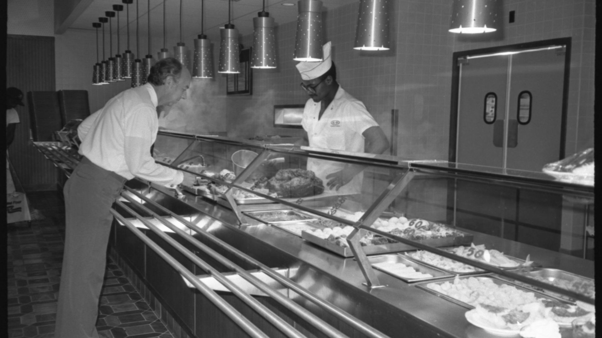 Black and white photo of a man serving himself food from a steam table in a cafeteria while a worker in a white uniform and hat stands behind the counter. Pendant lights hang above.