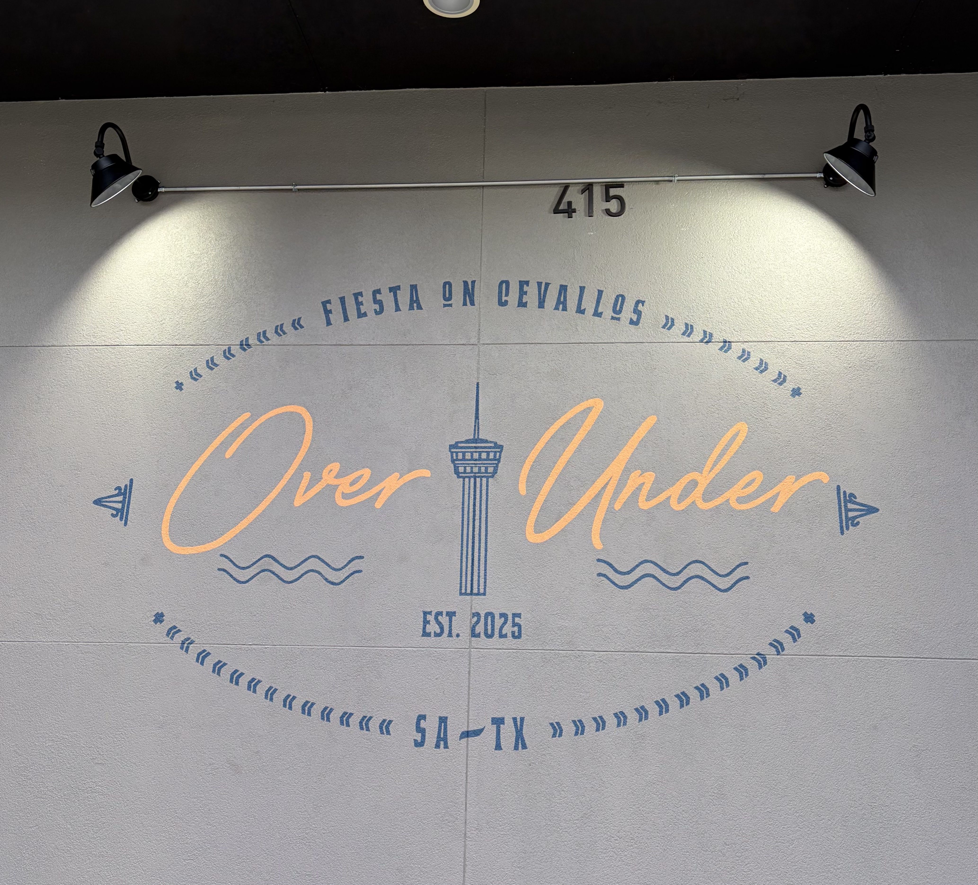 Wall sign with lights above reading "Fiesta on Cevallos Over Under Est. 2025" with San Antonio Tower icon and "SA-TX" below on a gray surface.