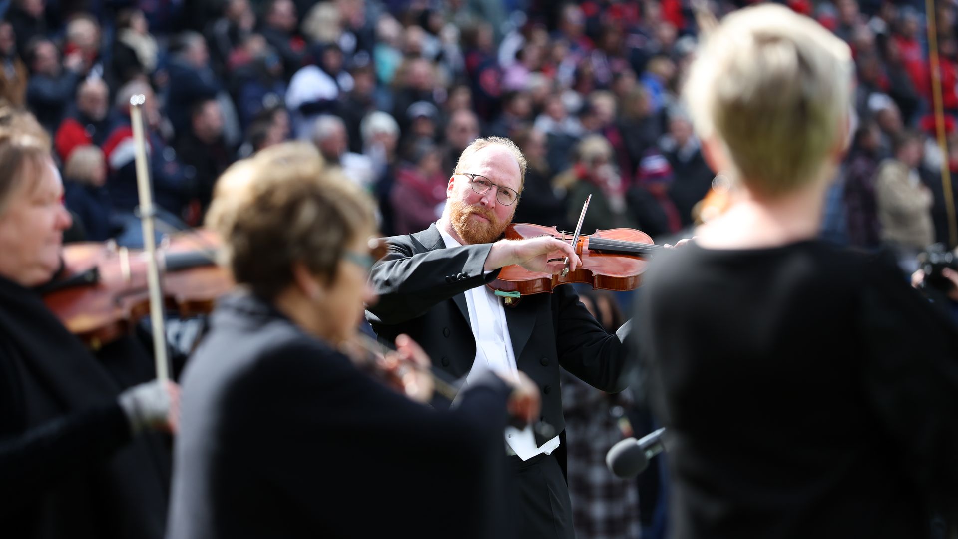 White man in tux playing violin in front of crowd; other instrumentalists out of focus.