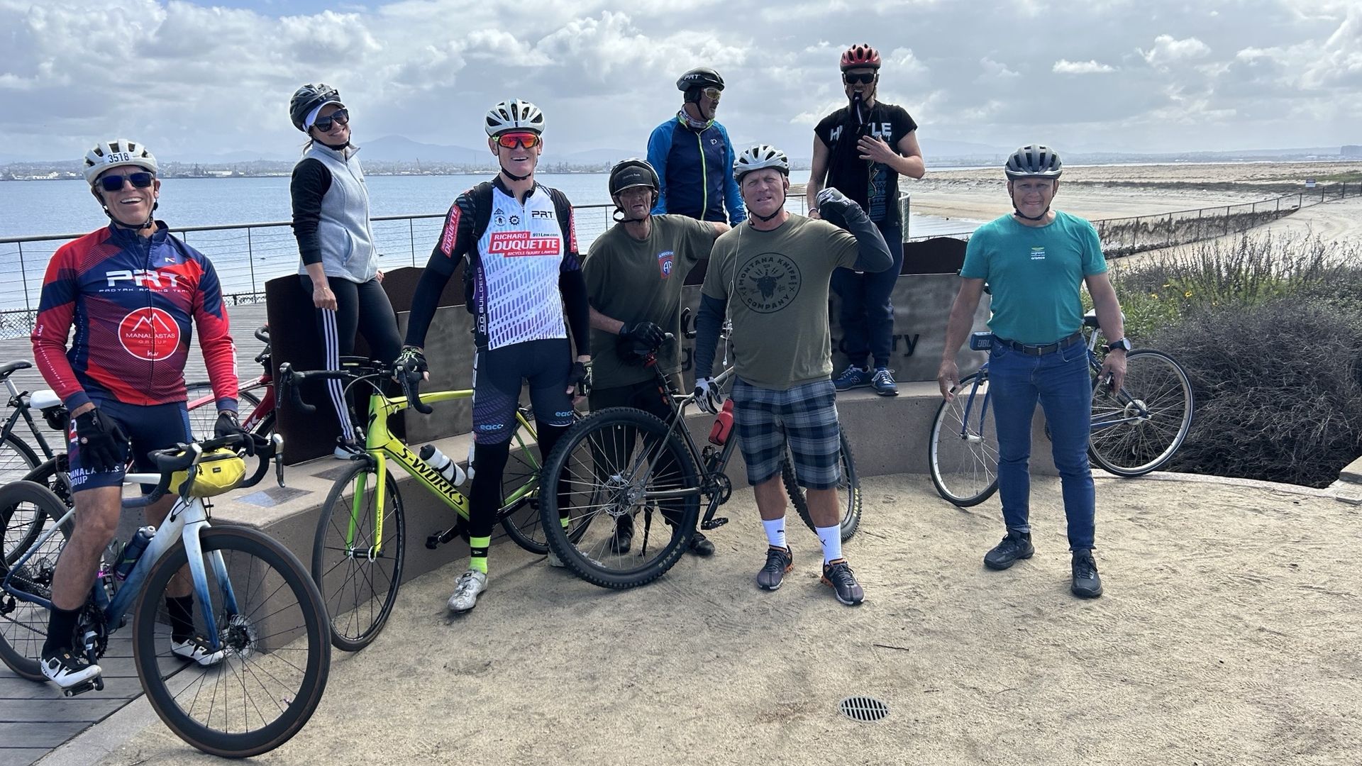 A group of eight people with bikes pose for a photo on the waterfront. 