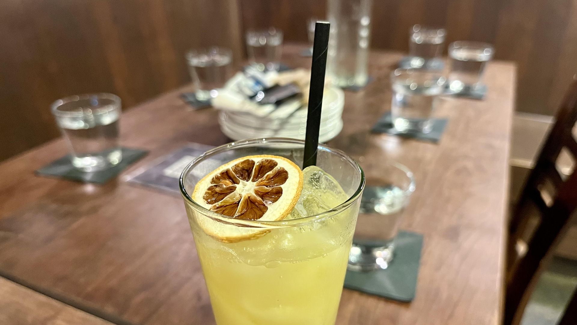 A yellow-colored drink in a tall glass with a dried citrus slice sits in front of a wooden table set with silverware and glasses.