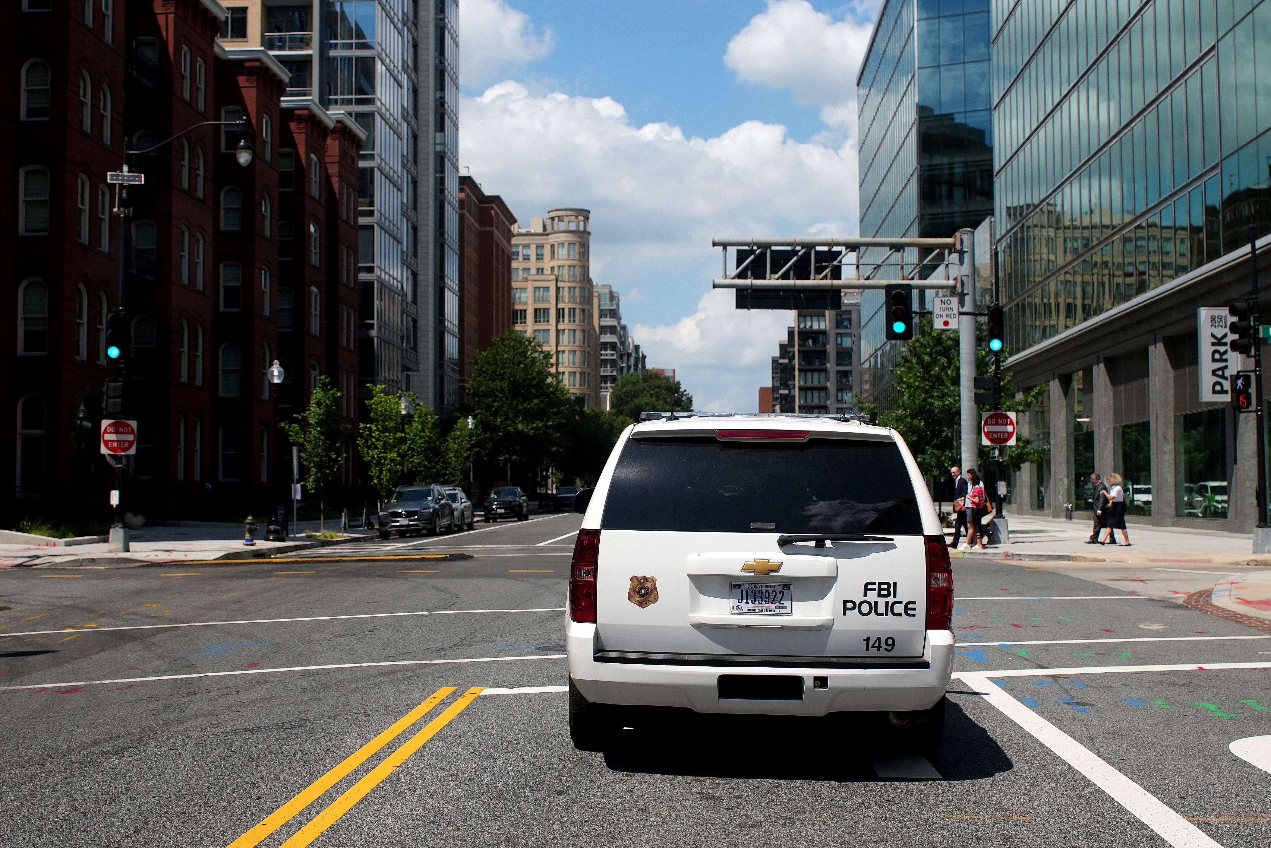 White FBI Police SUV with badge and number 149 at an urban intersection with traffic lights, tall buildings, pedestrians crossing, and a partly cloudy blue sky.