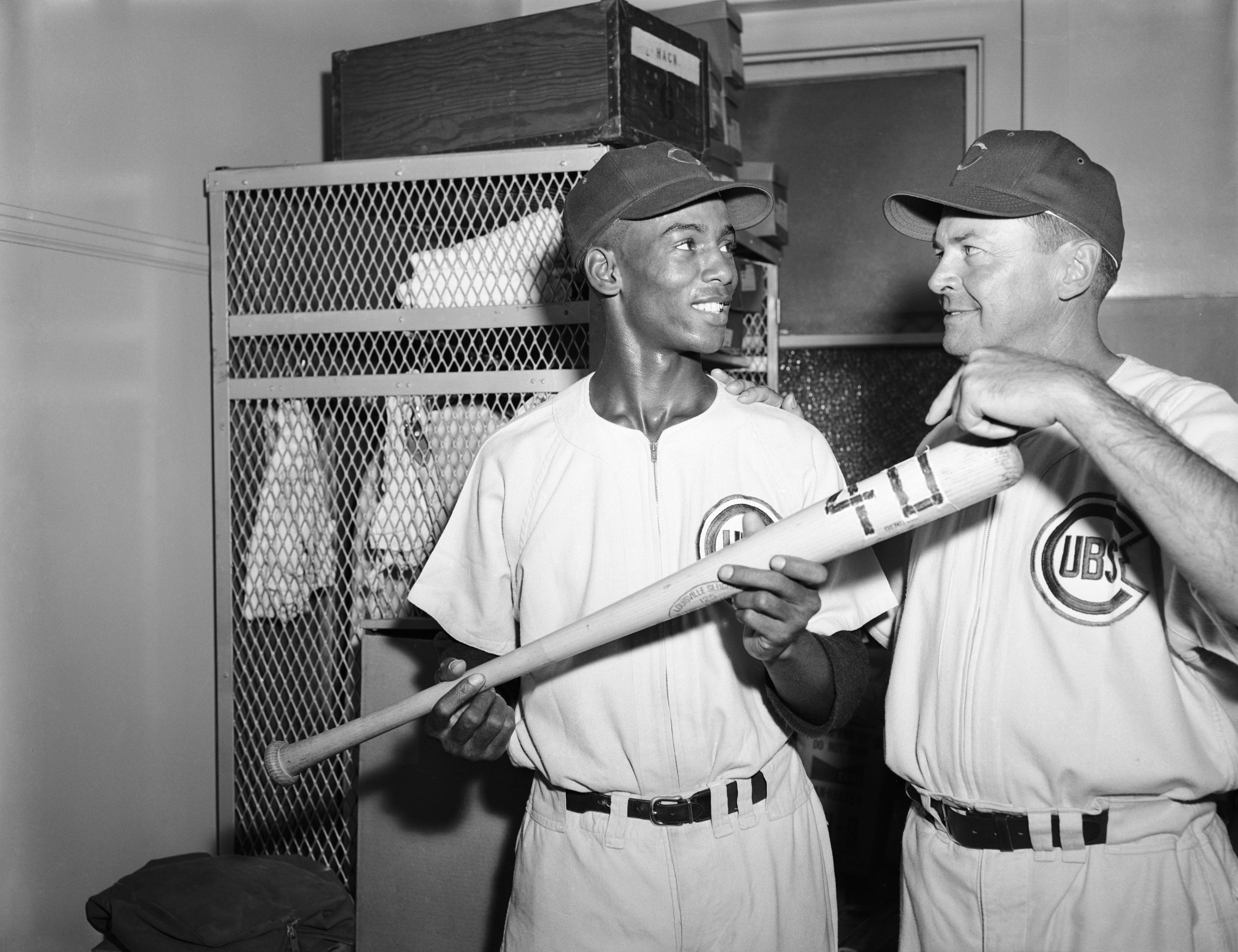 Photo of two people in baseball uniforms posing in front of a closet with a bat. 