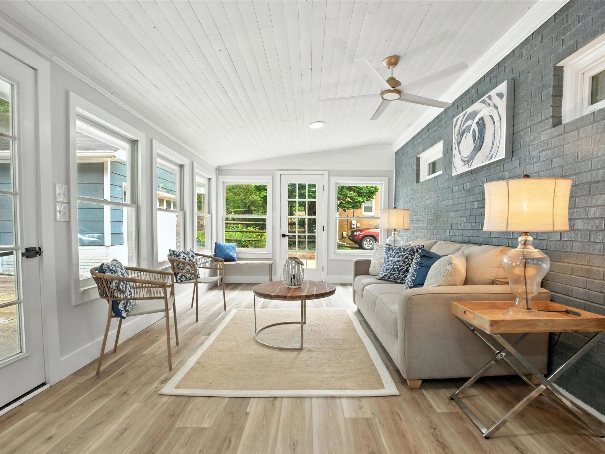 Bright sunroom with white ceiling and walls, gray brick accent wall, beige sofa with blue and white pillows, two modern chairs, wooden coffee table on beige rug, and lamps on side tables.