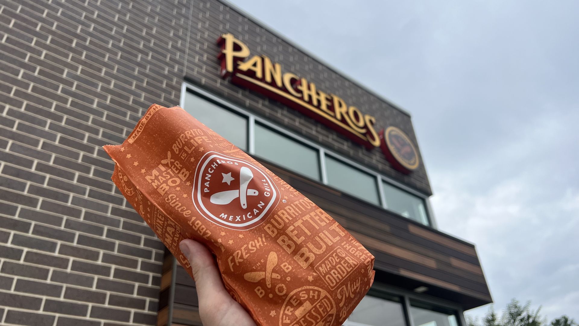 Close-up of an orange burrito wrapper with a Pancheros logo, held up in front of a Pancheros Mexican Grill storefront with a brown brick wall and blue sky.