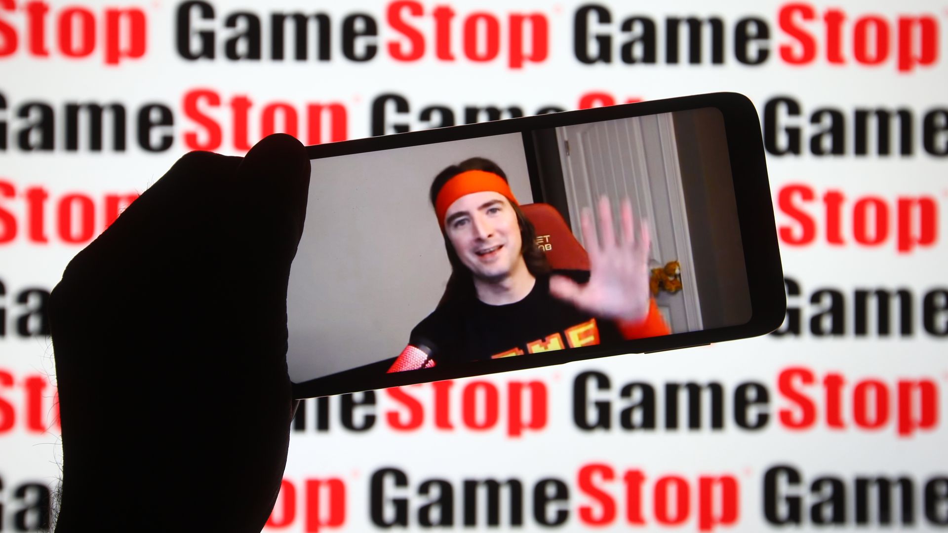 Keith Gill, aka Roaring Kitty, displayed on a smartphone with a GameStop sign in the backgrouund.