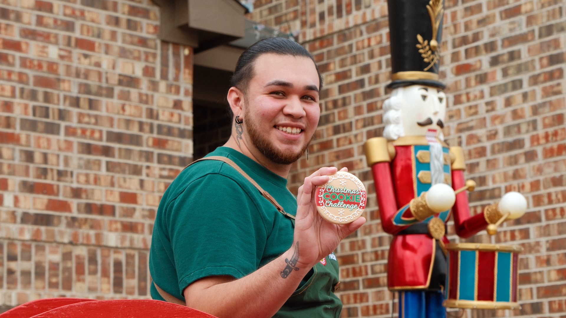Nelson Martinez, a Dallas baker, holds a cookie with the Food Network Christmas Cookie competition logo written in fondant. He is sitting in front of a decorative Christmas lawn ornament.