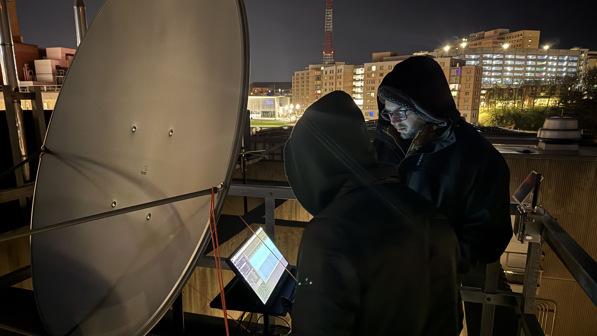 Two people in hooded jackets on a rooftop at night, working with a large satellite dish and a laptop. City lights, buildings, and a tall red-lit tower glow in the background.
