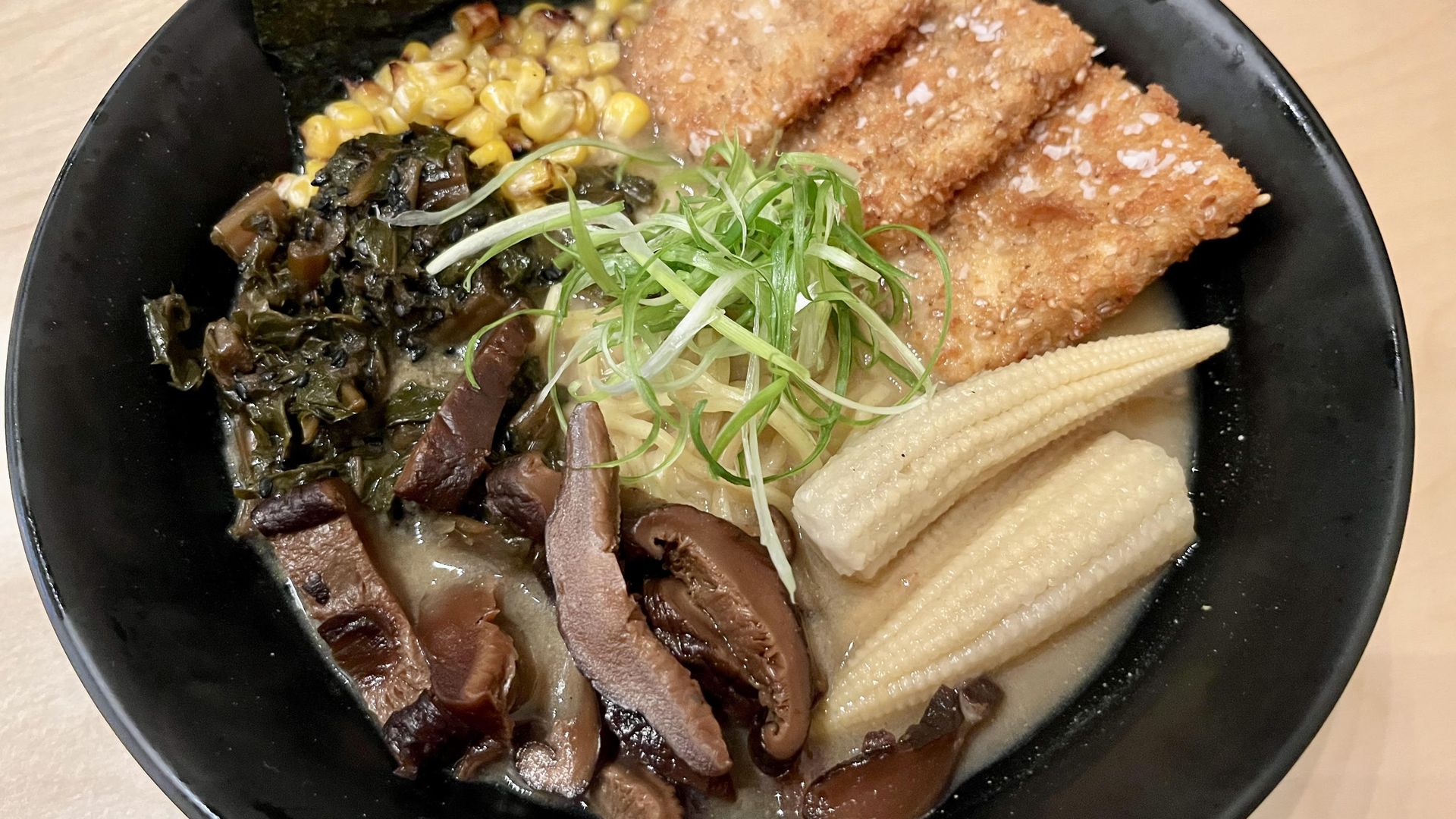 A large black bowl with ramen noodles topped with breaded fried slices of tofu, baby corn, grilled corn, mushrooms, green onions and seaweed all atop a creamy broth.