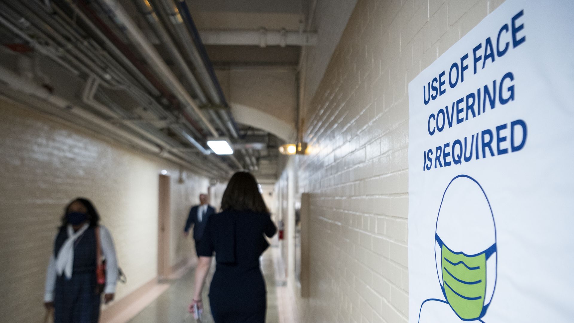 Photo of two people walking in a hallway as a sign that says "Use of face covering required" is taped to a wall