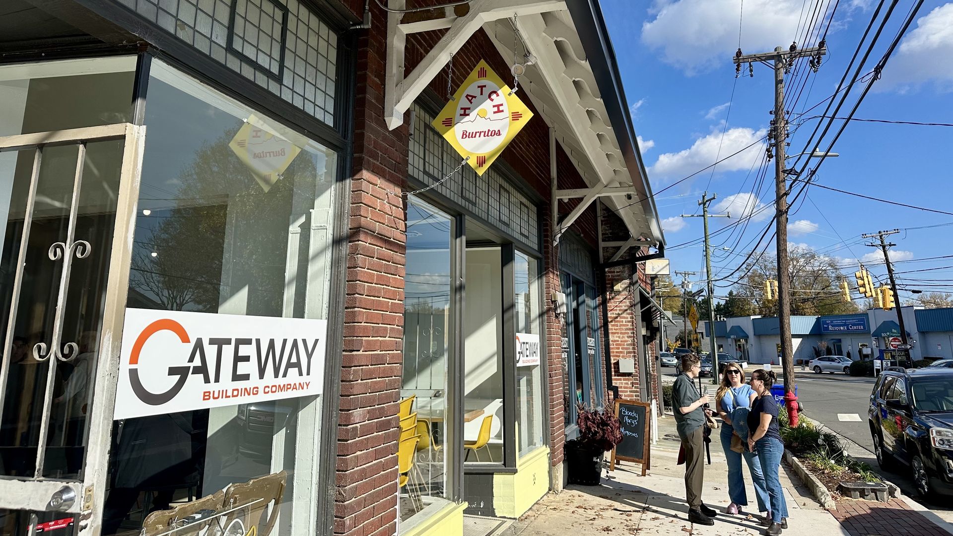 Brick building with signs for "Gateway Building Company" and Hatch Burritos. Three people stand on the sidewalk under a blue sky with clouds and power lines overhead.