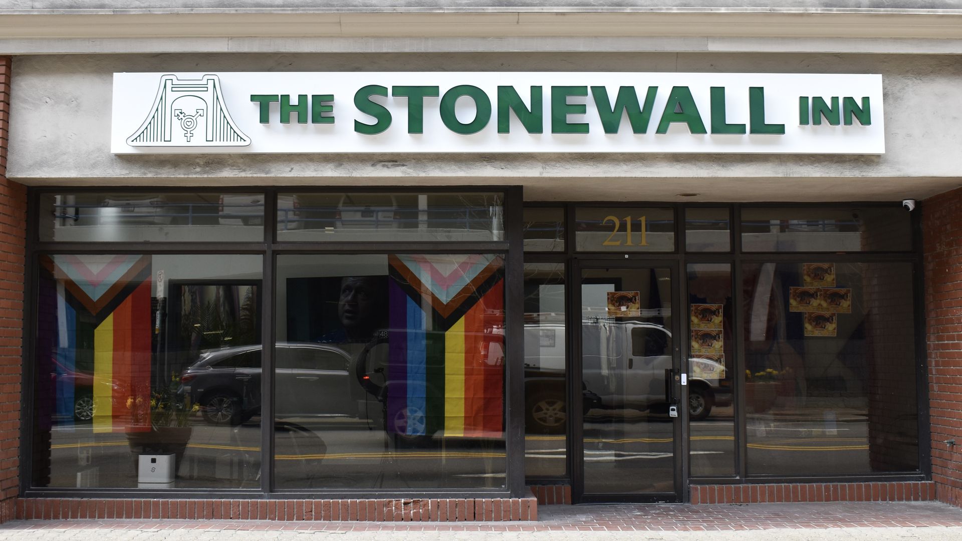 Storefront of "The Stonewall Inn" with a white sign and bold green letters, a bridge logo on the left. Large glass windows show rainbow Pride banners inside; brick base and street reflection.