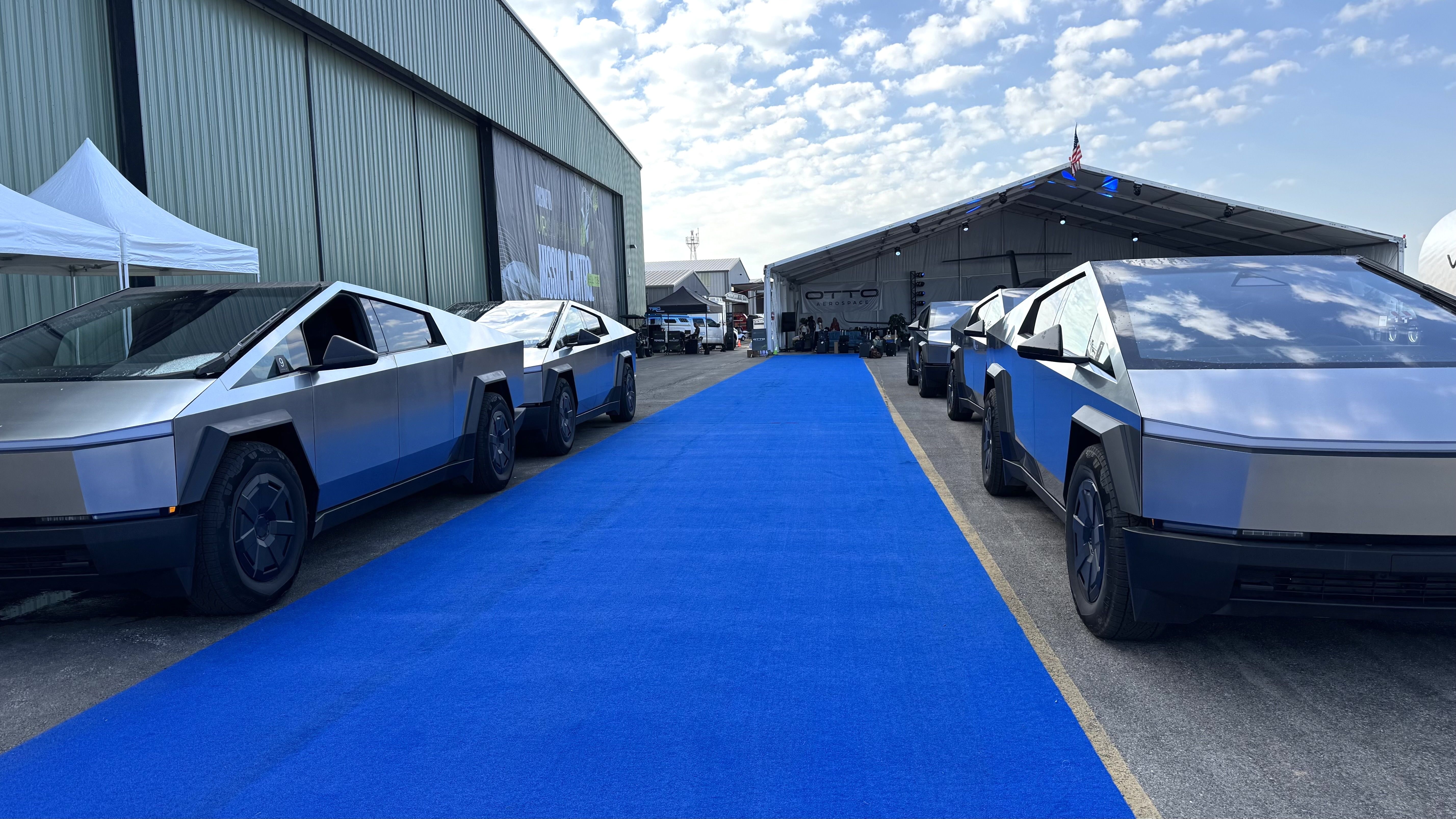 Several Tesla Cybertrucks are parked on either side of a bright blue carpet leading to a large event tent at an airfield. The vehicles are lined up in front of a green hangar under a partly cloudy sky.
