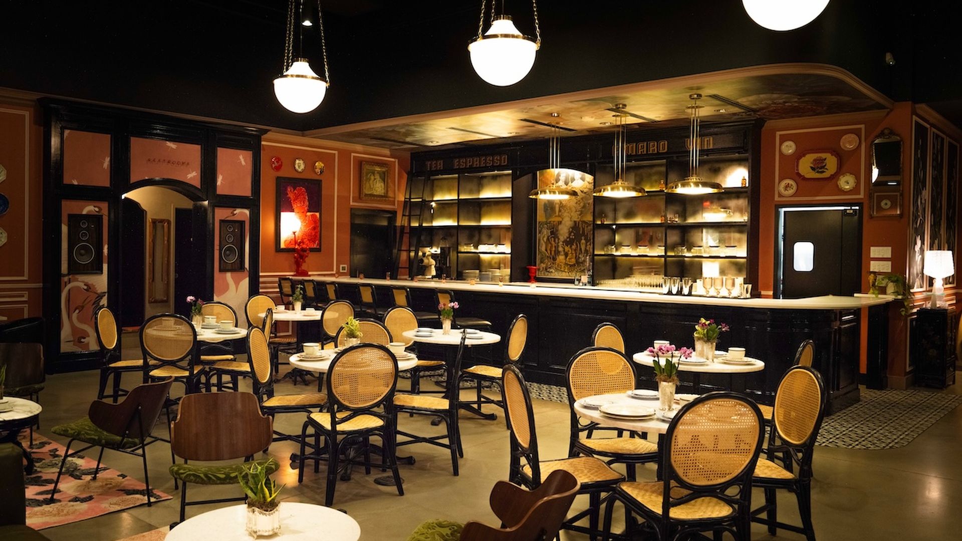 Retro cafe interior with round white tables, wicker seating, and a long black bar. Warm pendant lights, orange walls with art, and small flower vases on tables create a cozy ambiance.