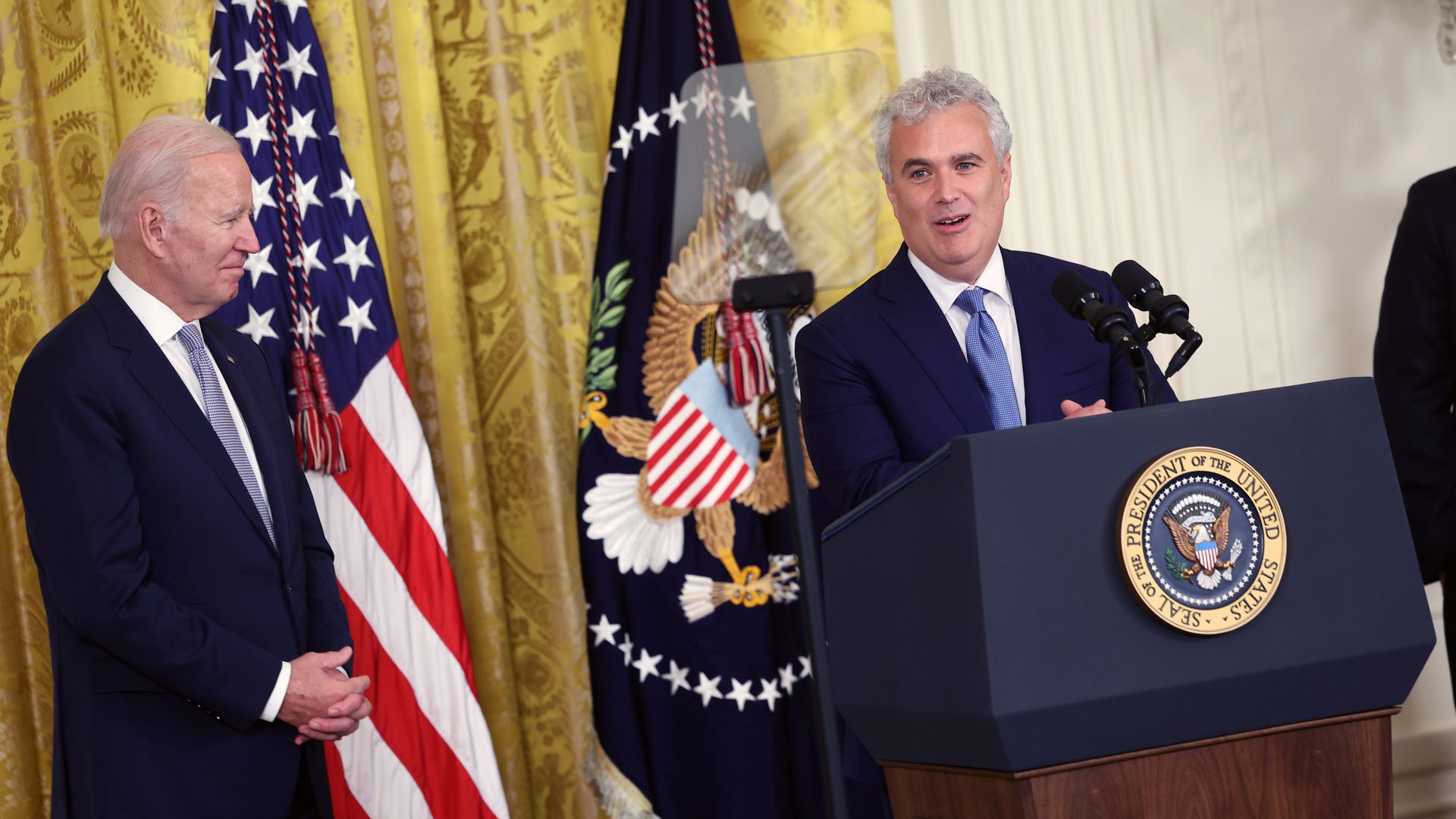 Joe Biden looks onto Jeff Zients before flags and the golden drapes of the east room of the White House