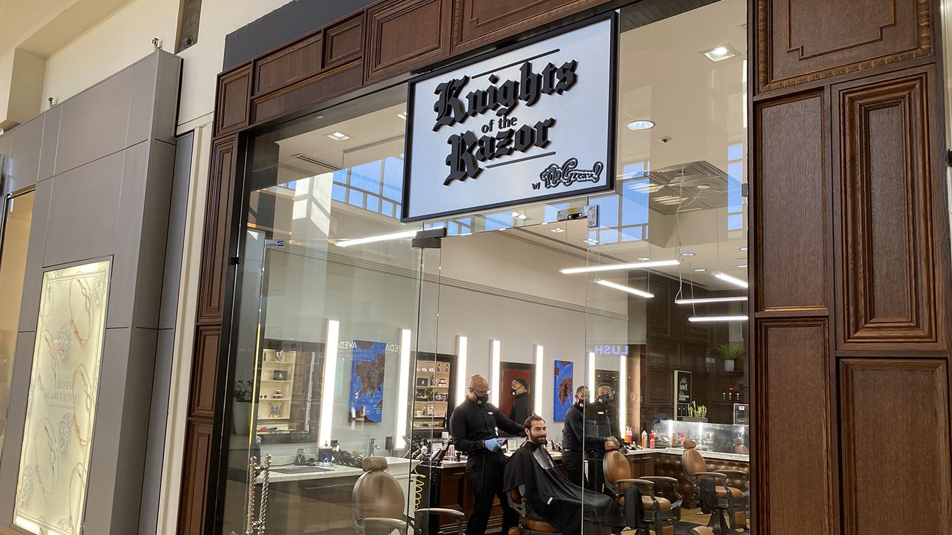 outside of a mall barbershop. glass opening surrounded by wood. "Knights of the Razor" sign over the door. A barber standing behind a customer in a chair