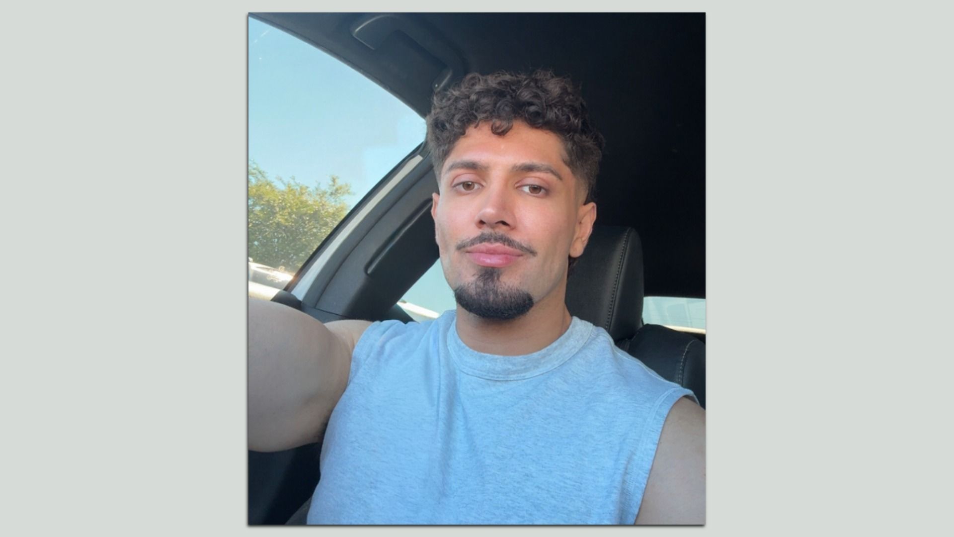 Man with curly hair and trimmed beard wearing a light blue sleeveless shirt, sitting in a car with trees and blue sky visible through the window.