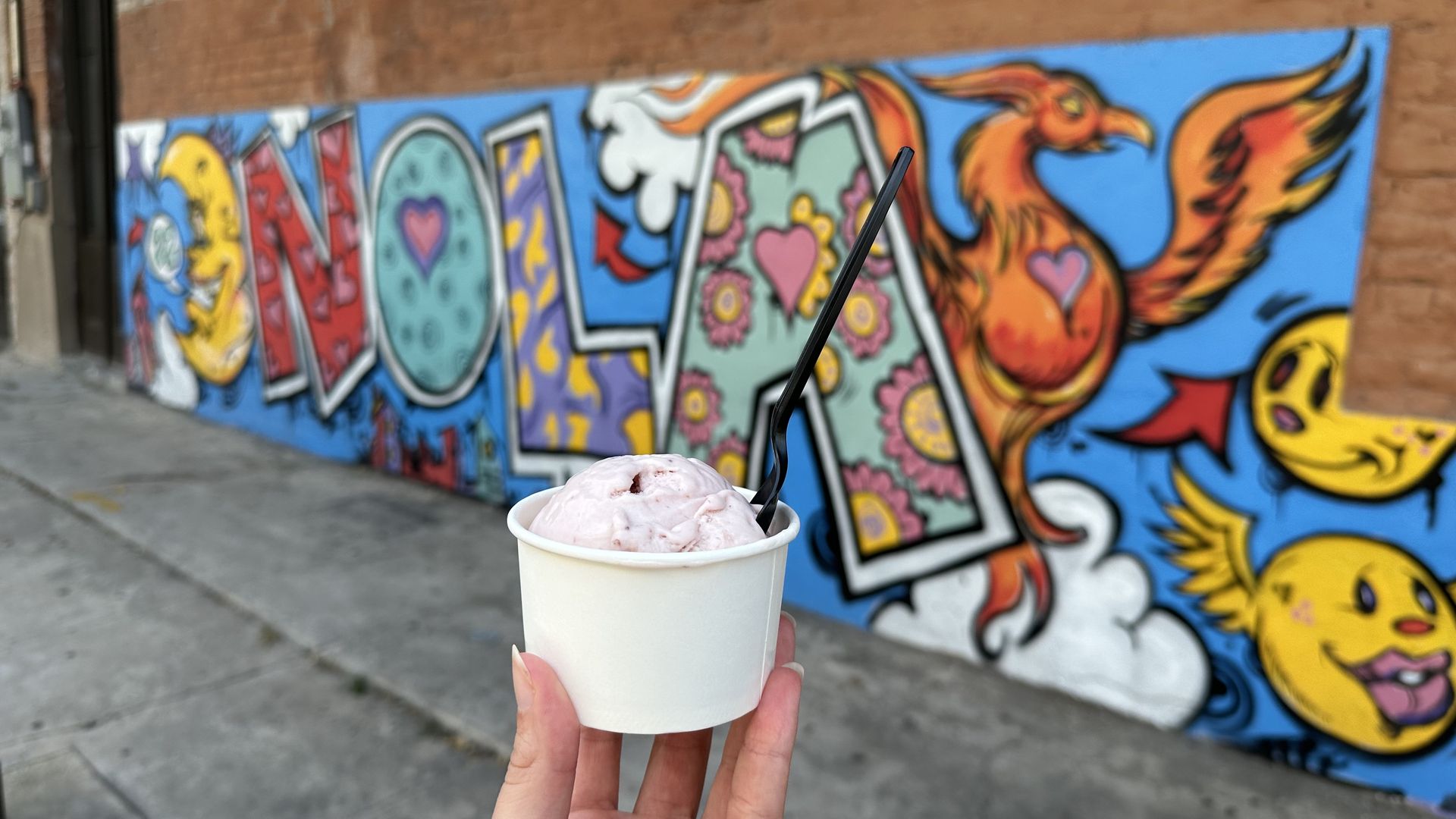 A cup of strawberry ice cream is held up in front of a colorful mural that says "NOLA." 
