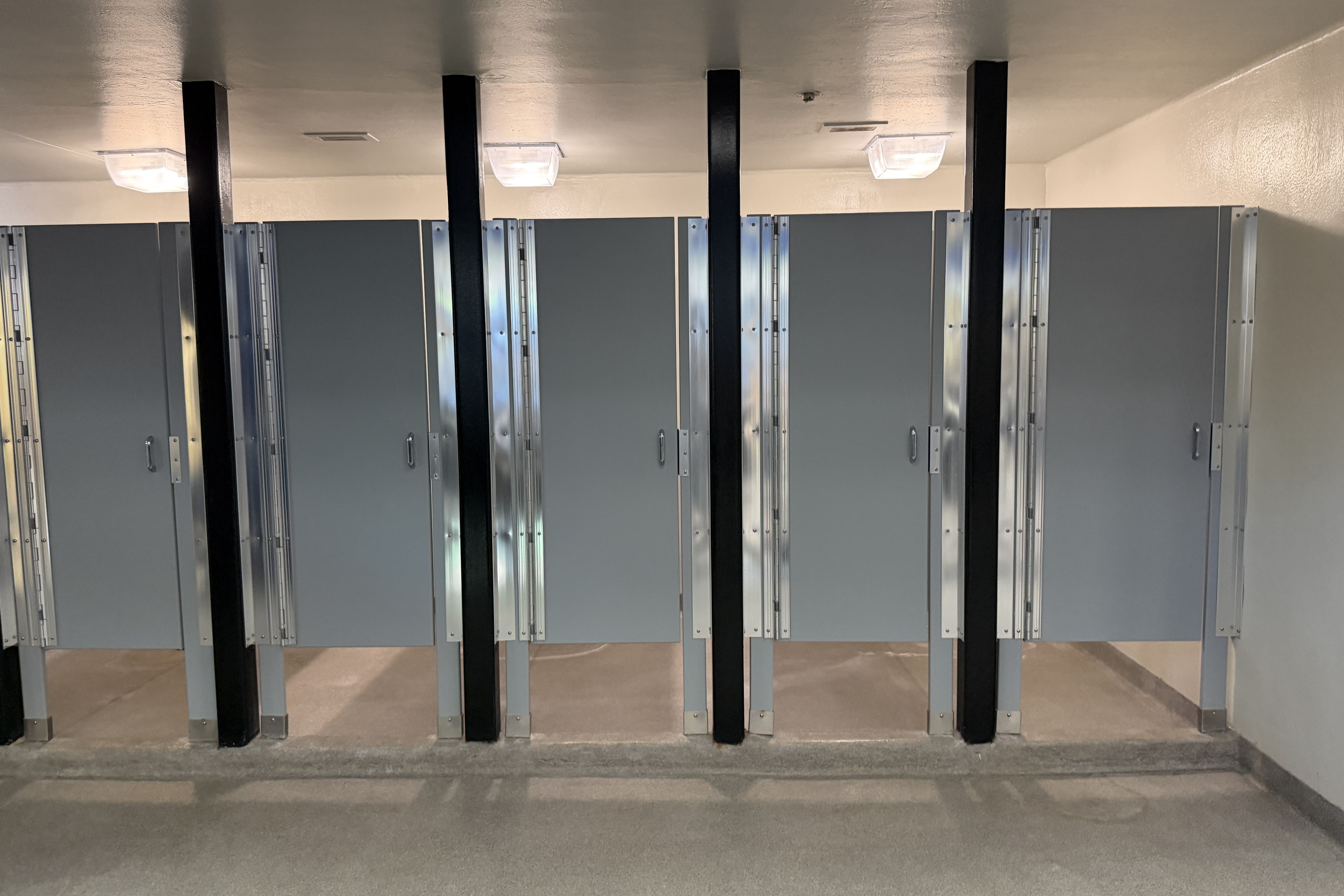 Row of gray metal stall doors with black vertical supports in a locker-room or public restroom; fluorescent ceiling lights, pale walls, and a concrete floor.