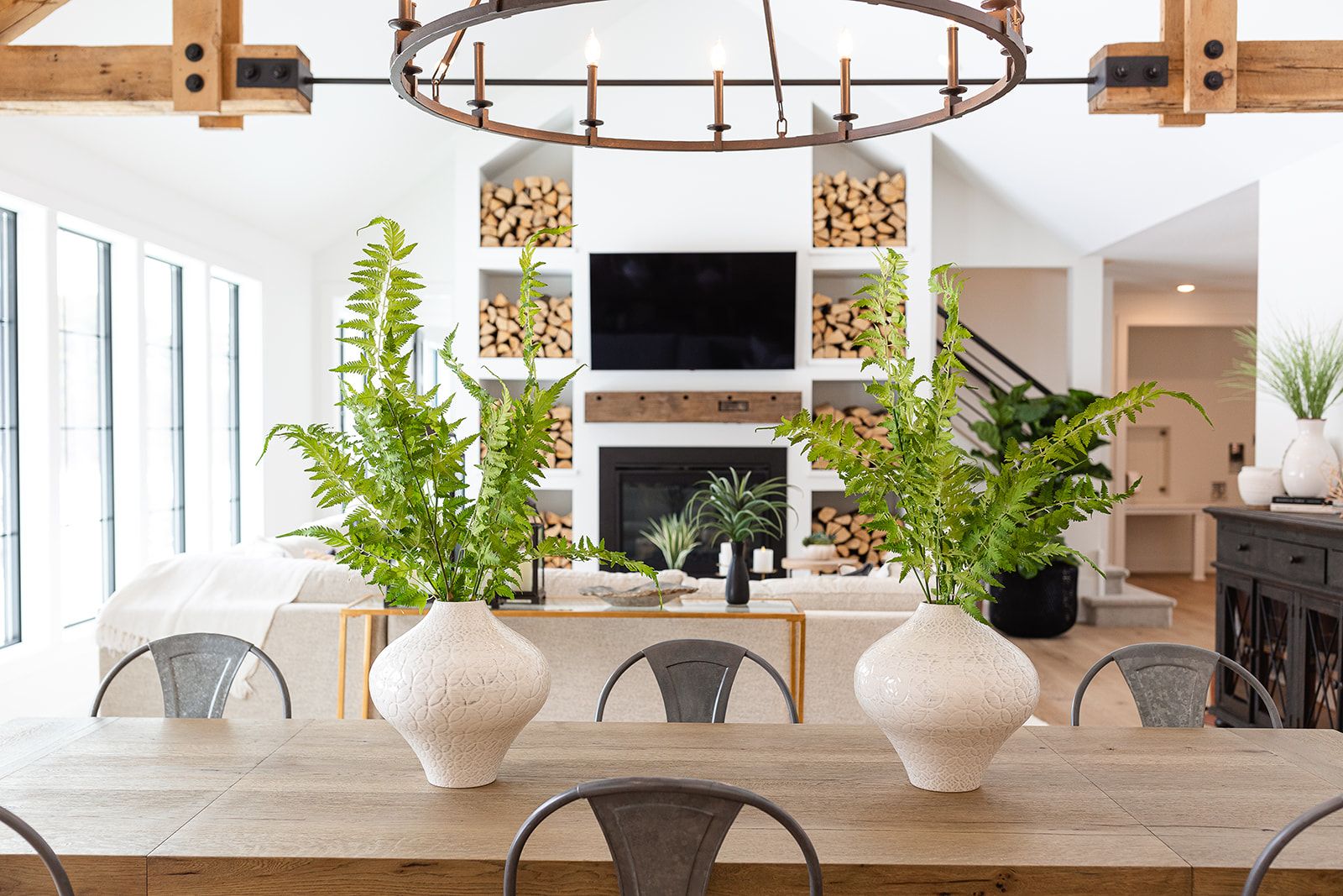 A photo showing the interior of a home with a fireplace flanked with shelving full of cut wood, metal dining chairs near an island and two plants as accents
