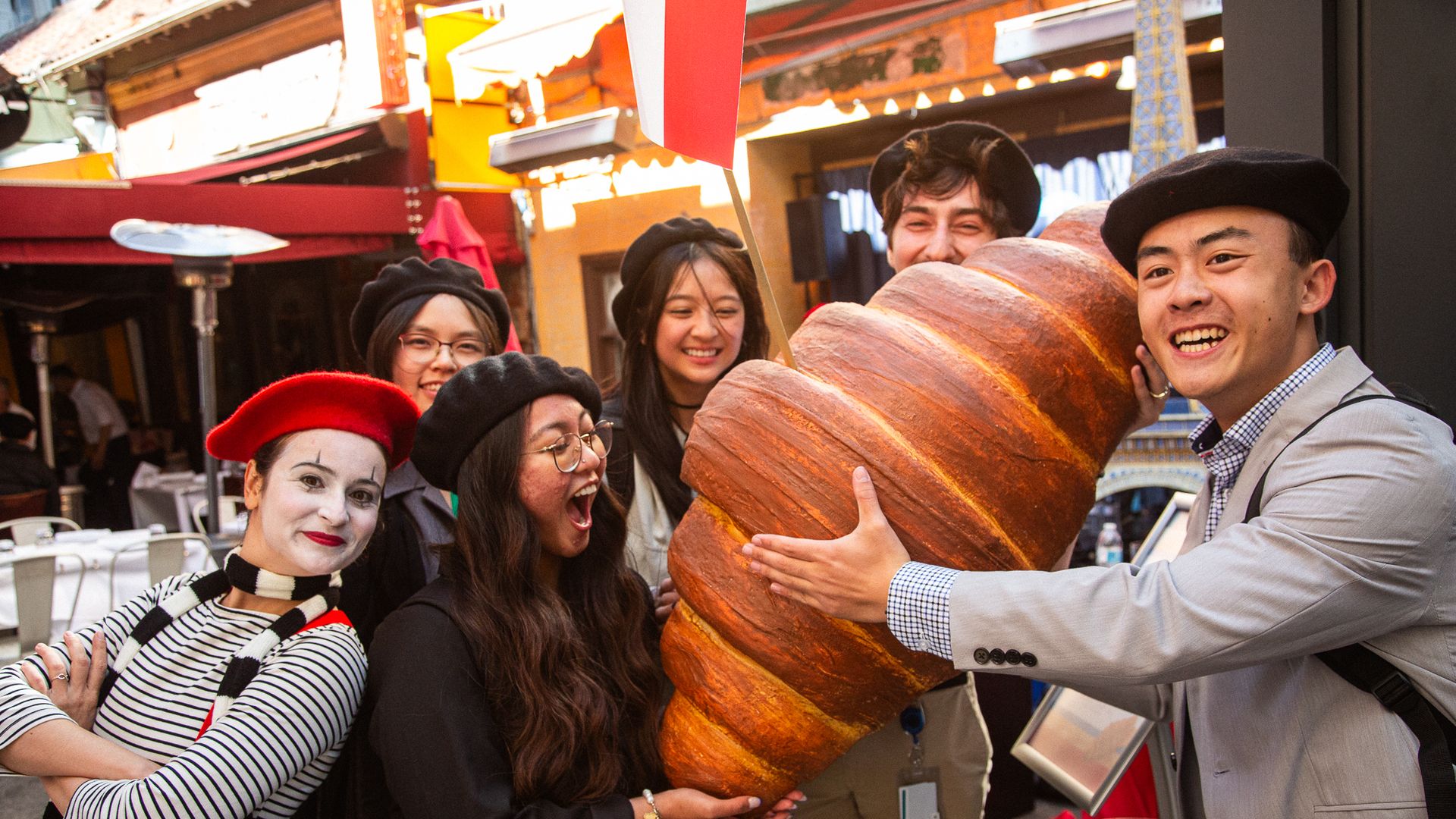A smiling group of friends posing with a giant croissant prop at a sunny outdoor market. They wear berets and striped outfits; a mime with white makeup stands in the foreground.