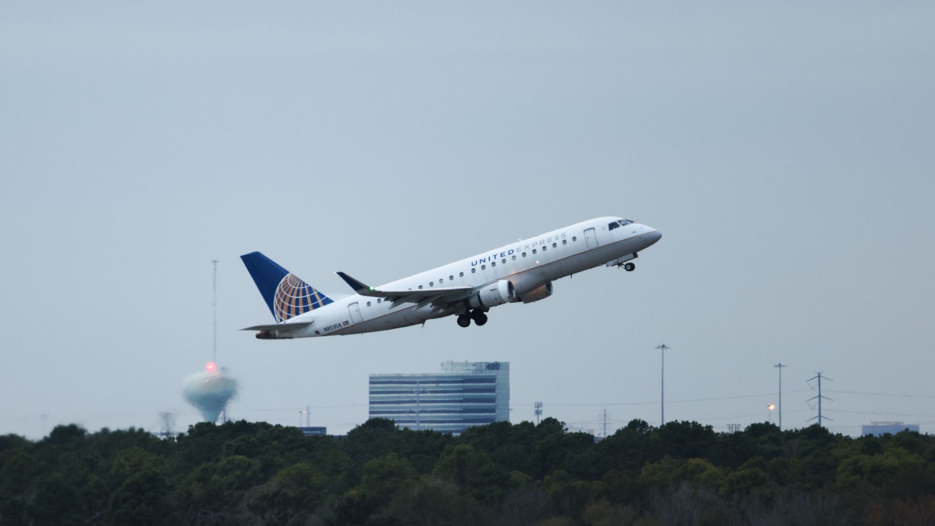 A United Express regional jet takes off, white fuselage and blue tail with a globe logo, climbing above a treeline with distant office buildings in the hazy sky.