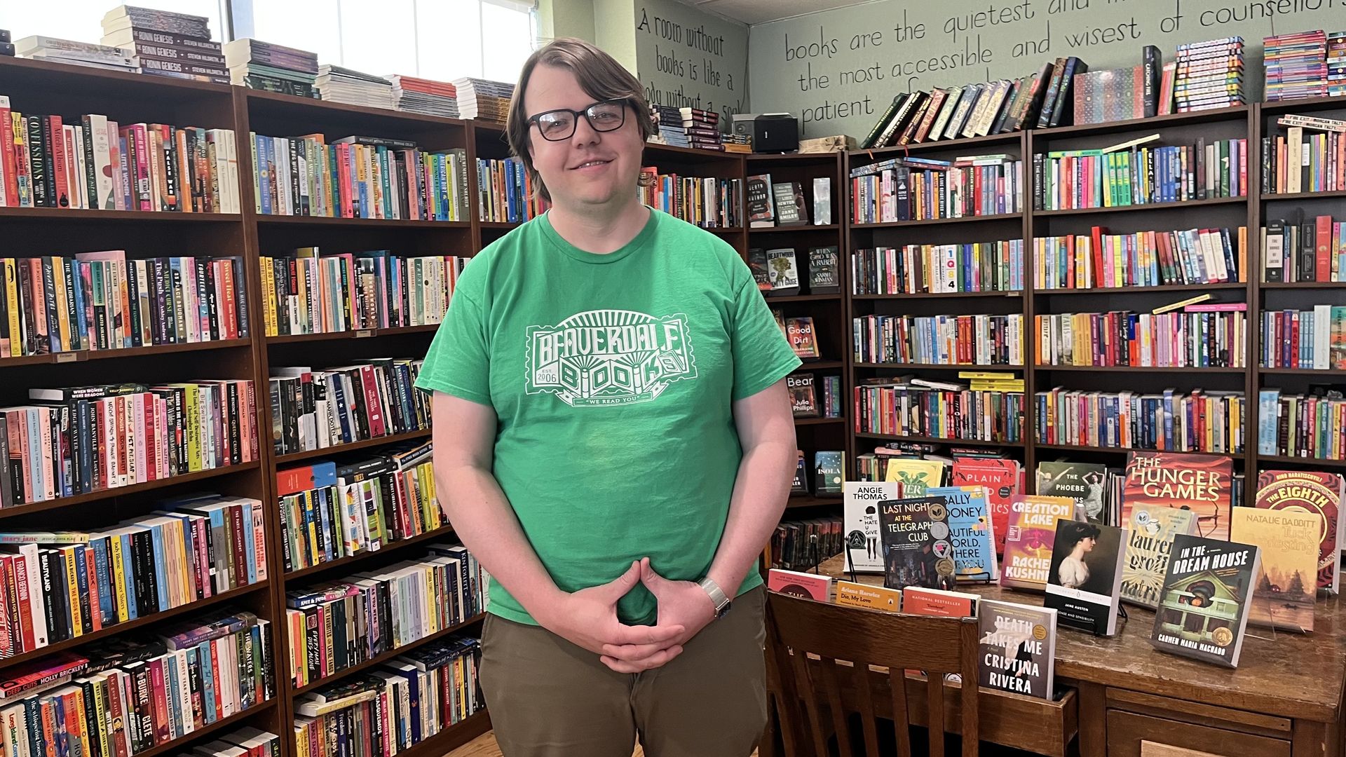 A person with short hair and glasses stands in a bookstore wearing a green t-shirt, smiling at the camera with hands clasped in front. Shelves of colorful books surround them, quotes on the wall.
