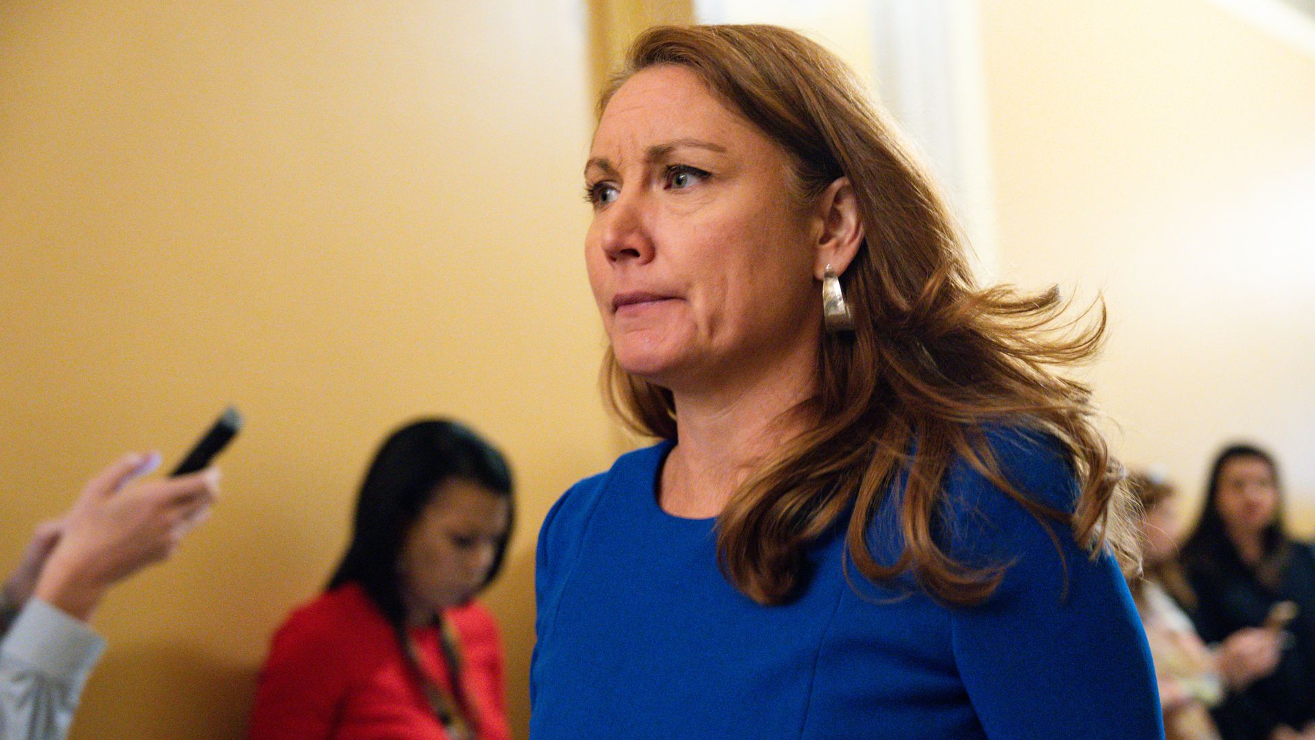Rep. Melanie Stansbury, wearing a blue dress, walks through a yellow hallway with reporters standing by the walls.