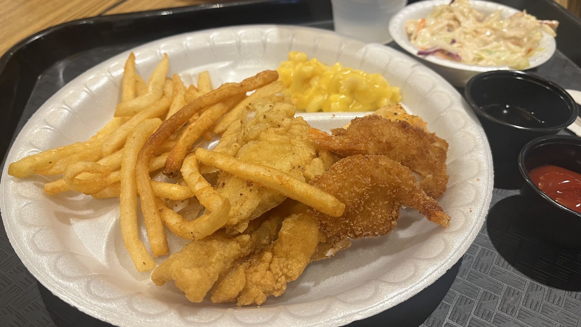 Foam plate with crispy fried chicken pieces, French fries, and macaroni and cheese; background shows coleslaw and small cups of ketchup and dark sauce.