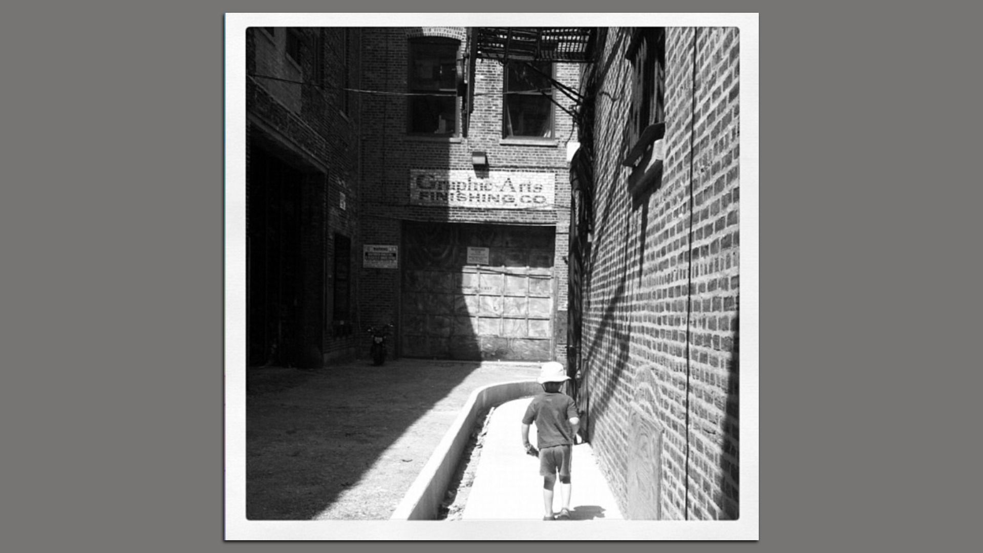 Black and white photo of a child wearing a hat walking along a narrow sunlit alley with brick walls, near a faded sign reading "Graphic Arts Finishing Co."