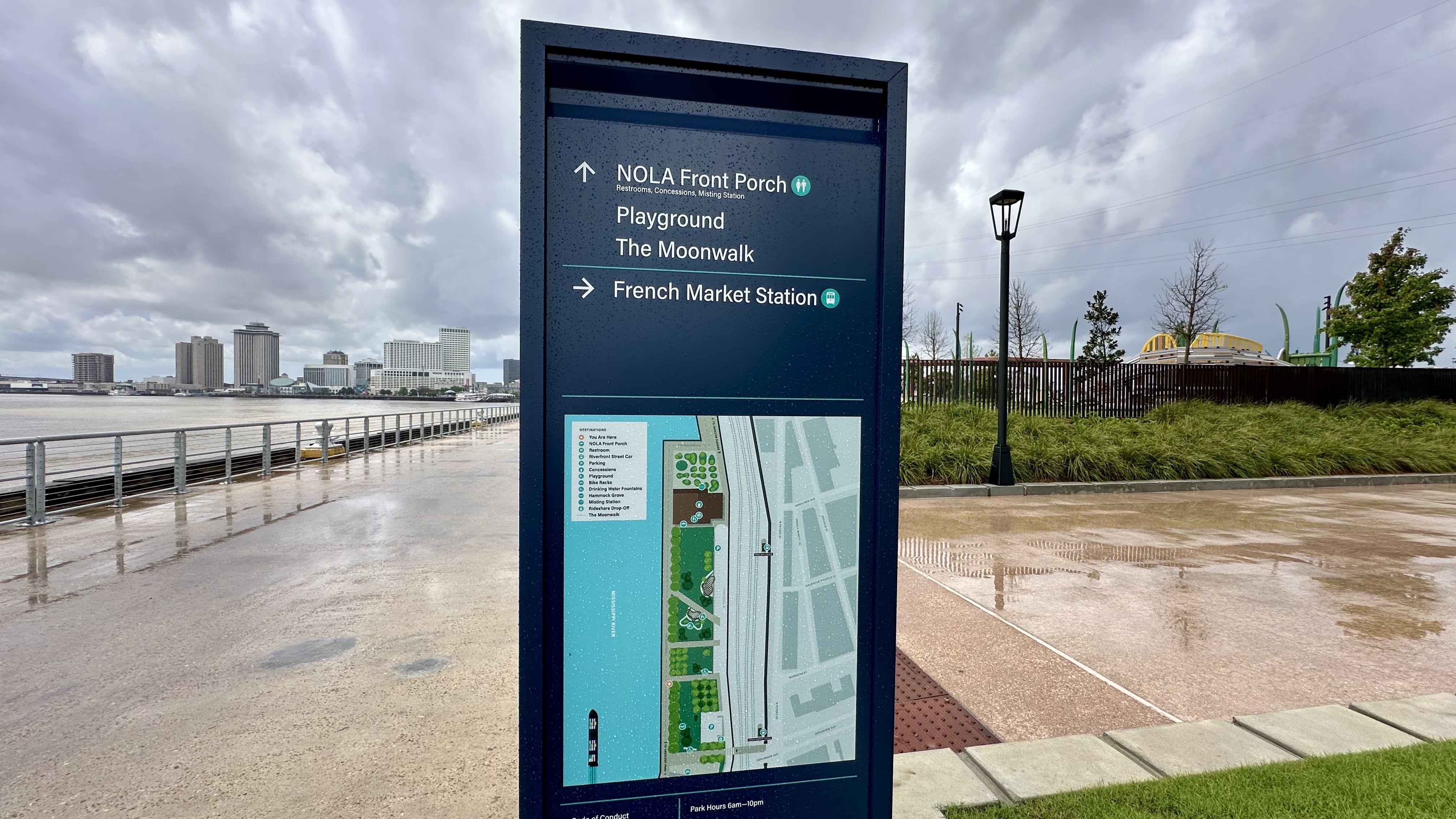 Blue riverfront sign with a map, directing to NOLA Front Porch, Playground "The Moonwalk", and French Market Station. Rainy day, wet pavement, railing along the water, distant city skyline.
