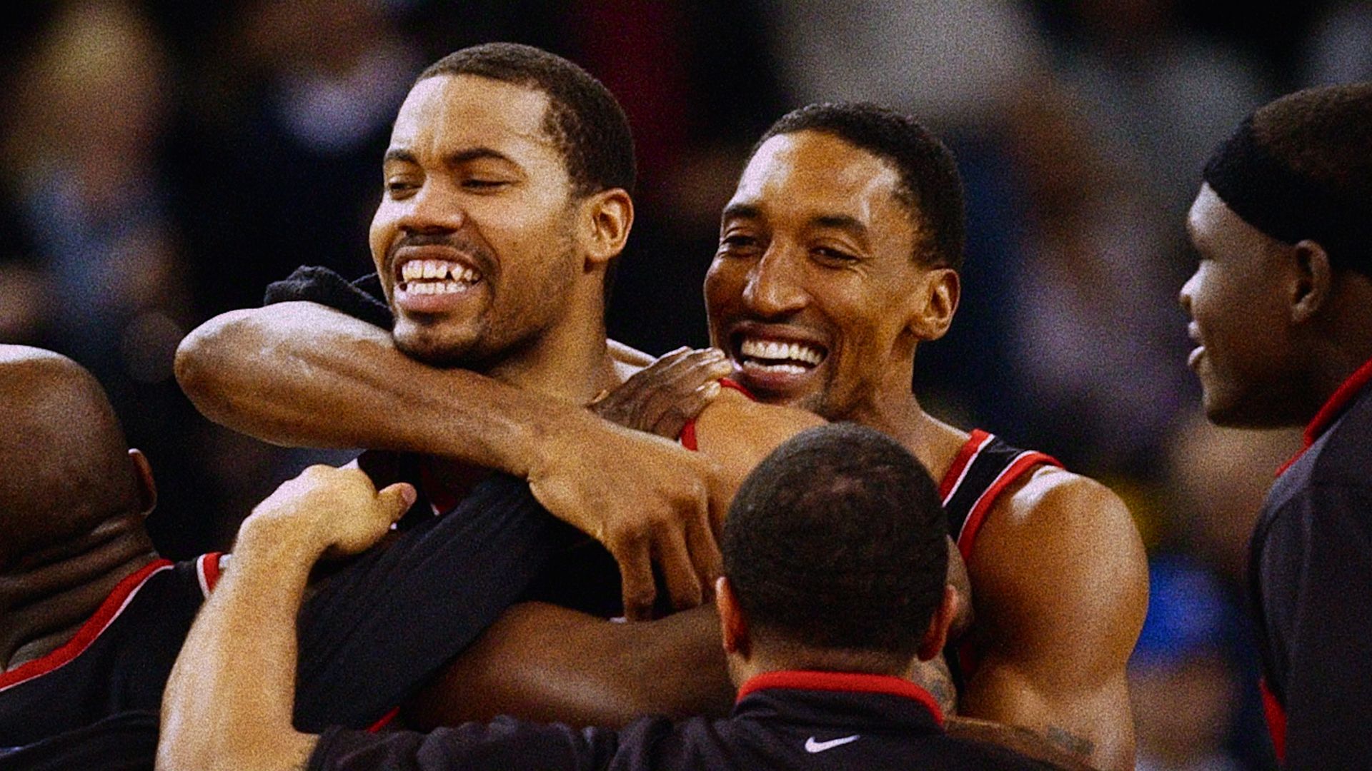 Basketball teammates in black uniforms with red trim embrace and celebrate a win, smiling broadly as they hug, with a blurred crowd in the background.