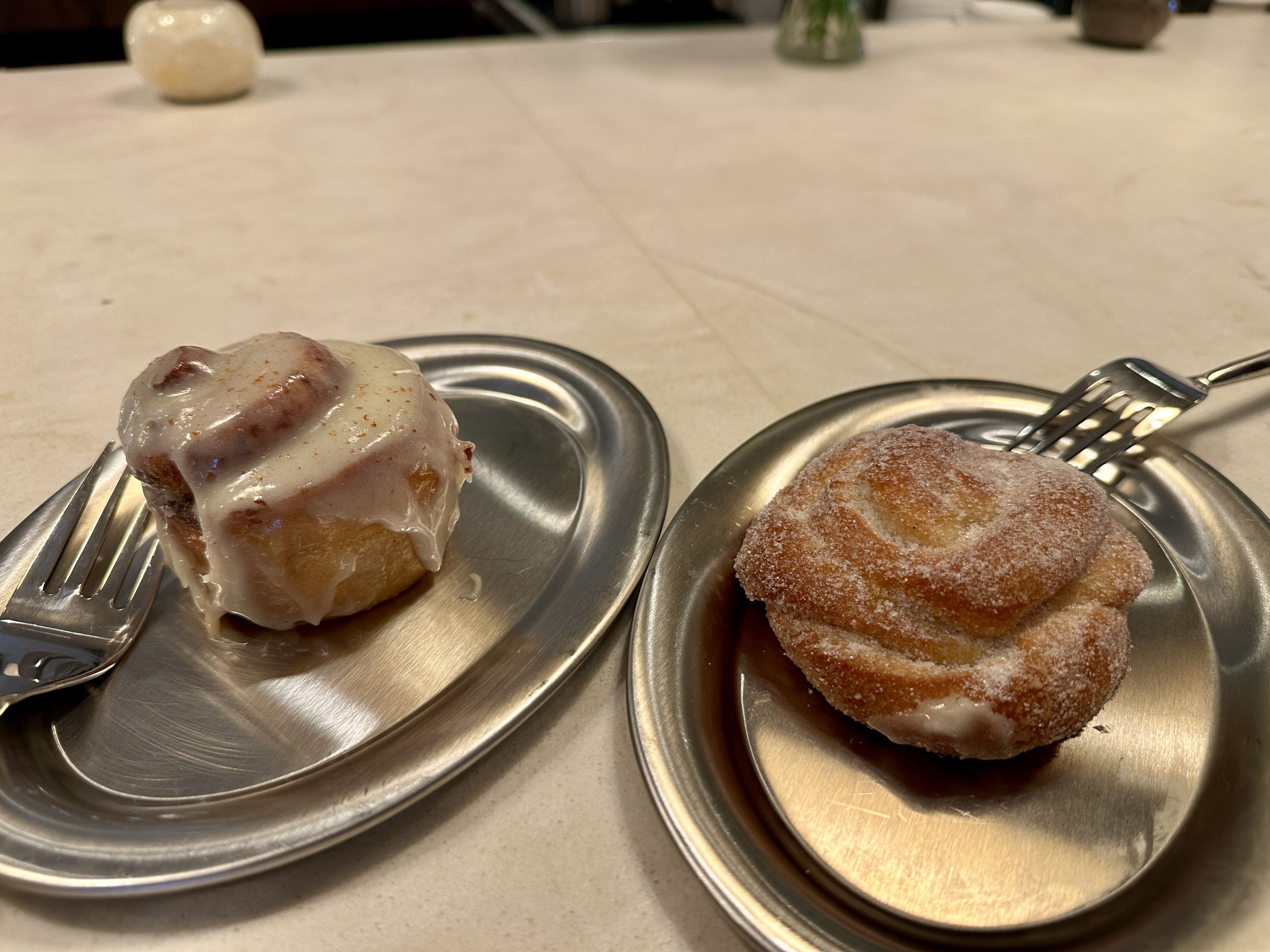 Close-up of two metal plates on a light table: left is a glazed cinnamon roll, right a stack of sugar-dusted donuts. Forks rest nearby, background shows a blurred shaker. HUckleberry Noda.