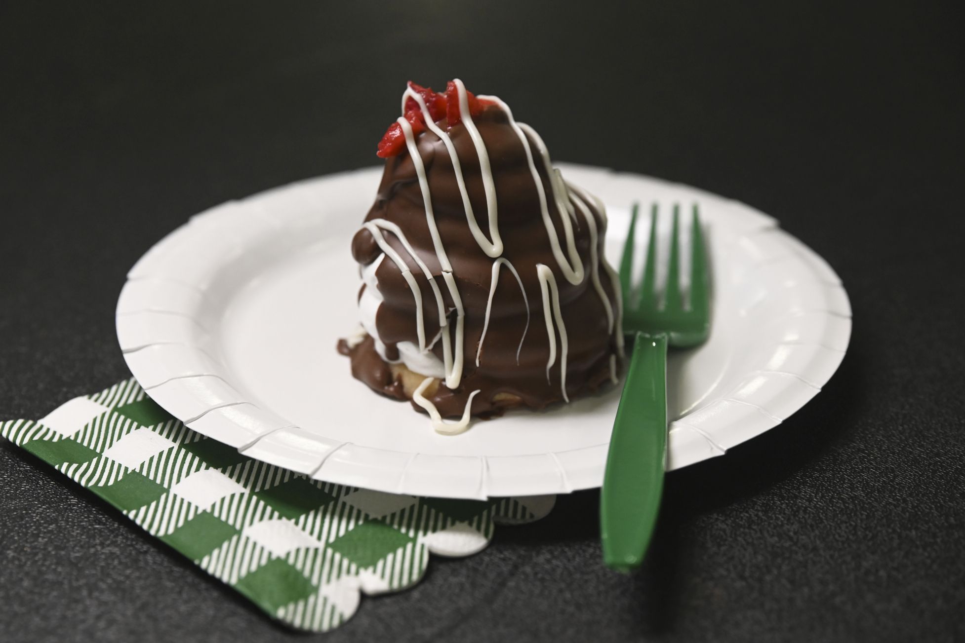 Chocolate-covered pastry with white icing drizzle and a red strawberry piece on a white paper plate; a green plastic fork and a green-and-white checkered napkin sit on a dark surface.