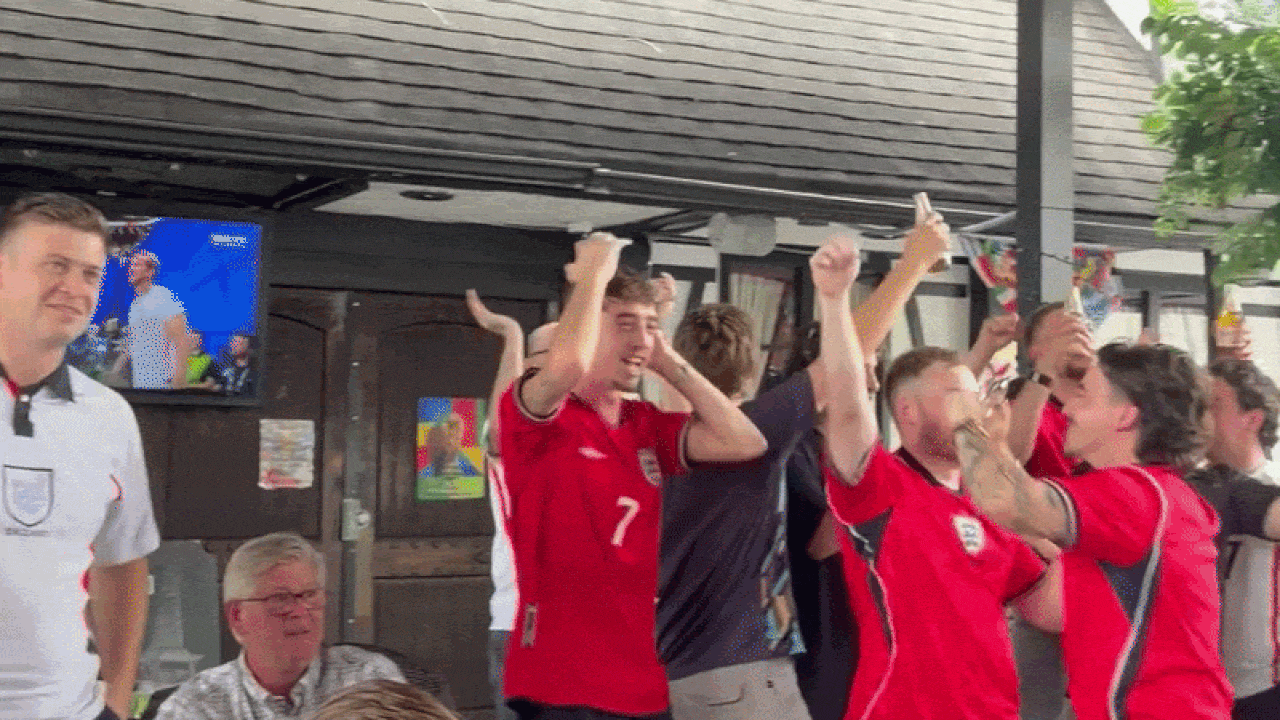 Group of men in red soccer jerseys cheer with arms raised in a bar-like venue; a white-clad official stands left as a seated man watches a celebrating crowd.
