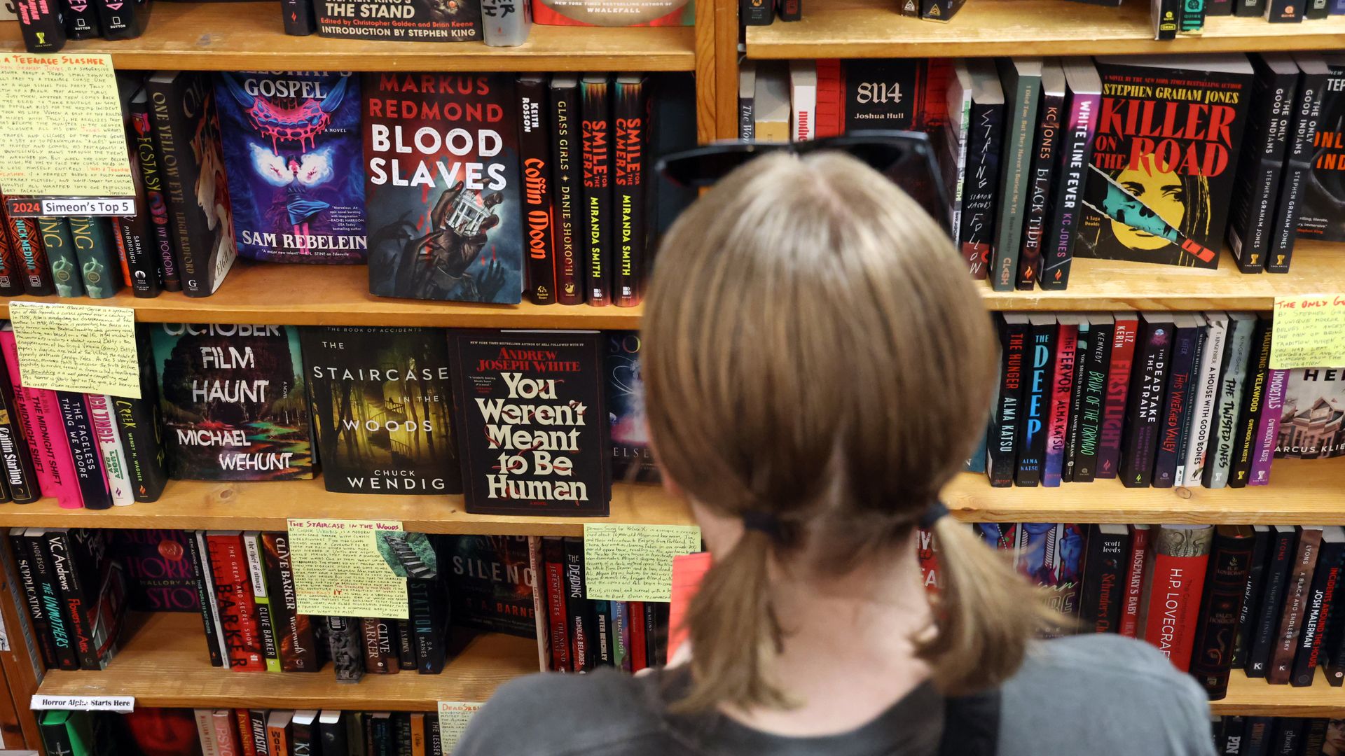 Back view of a person with light hair browsing crowded bookstore shelves filled with colorful thriller and horror novels, including a prominent "Blood Slaves" cover.
