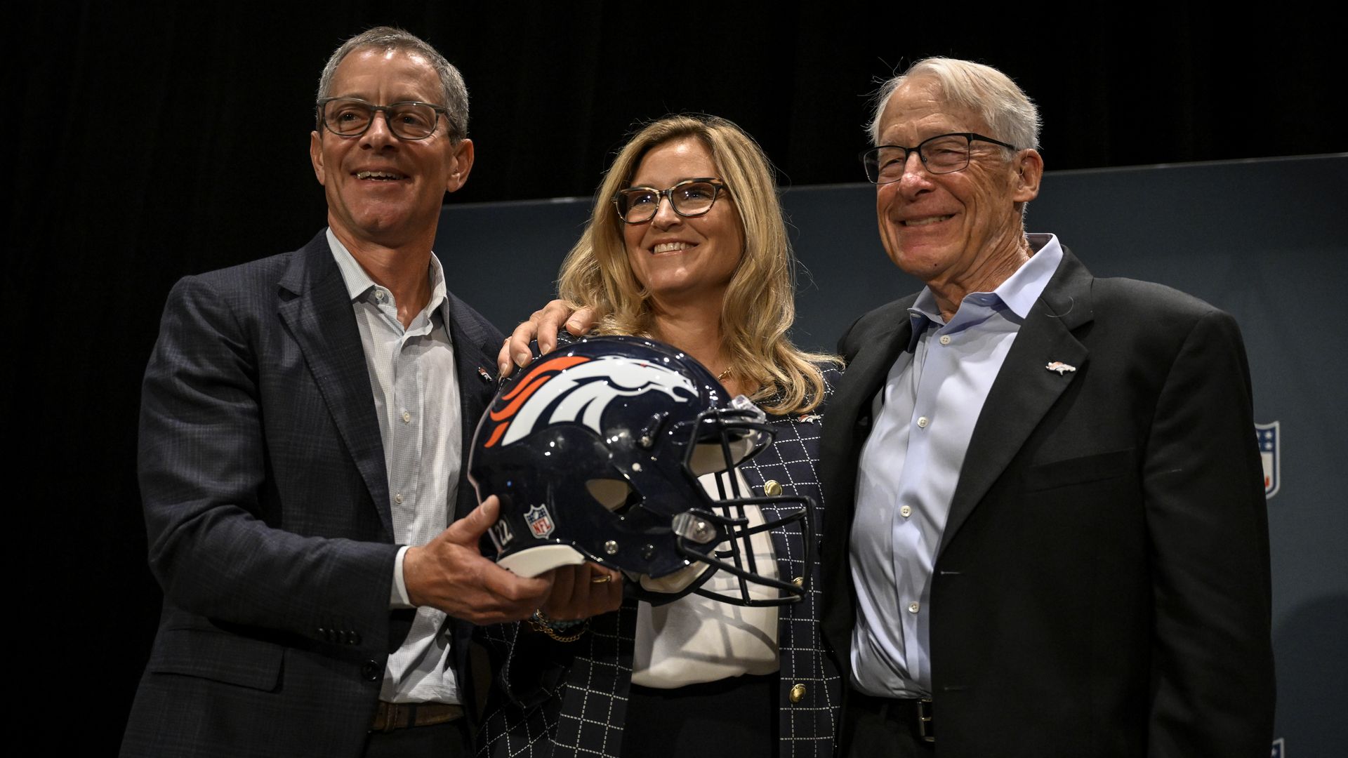Three people on a stage posing, a man hands a Denver Broncos helmet to a smiling blonde woman in a checkered blazer, with a suited man beside them; NFL logos in the backdrop.