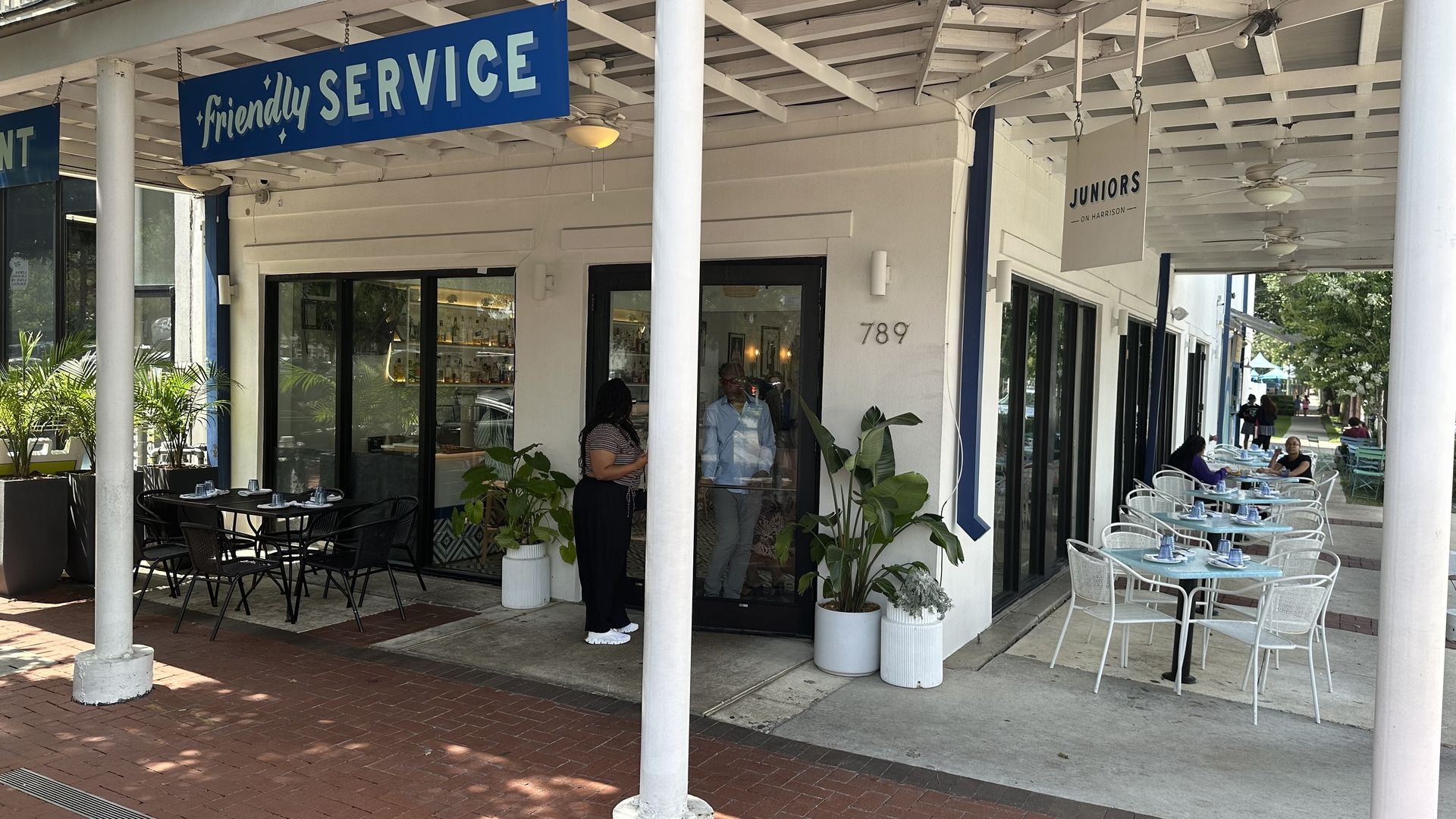 Outdoor view of a cafe with blue signs "friendly SERVICE" and "JUNIORS ON HARRISON", black and white chairs, blue tables set with napkins, and people seated and standing near the entrance.