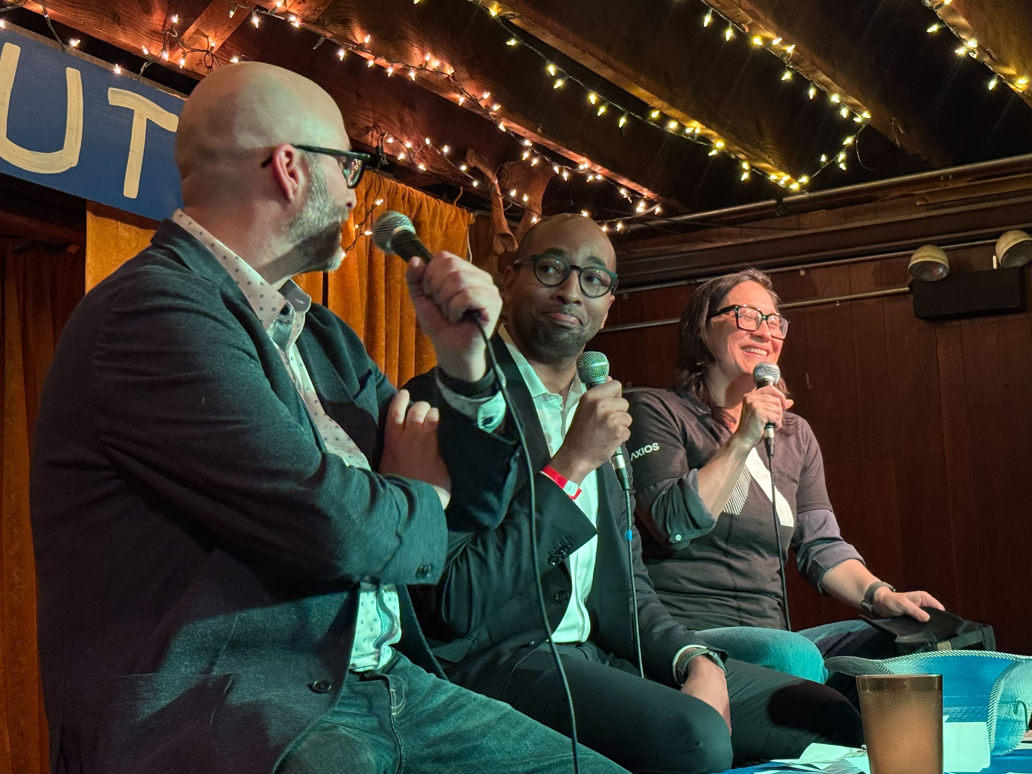 Three panelists on a cozy stage under string lights: a bald man in a blazer left, a man with round glasses center, and a smiling woman with glasses right, all holding microphones.