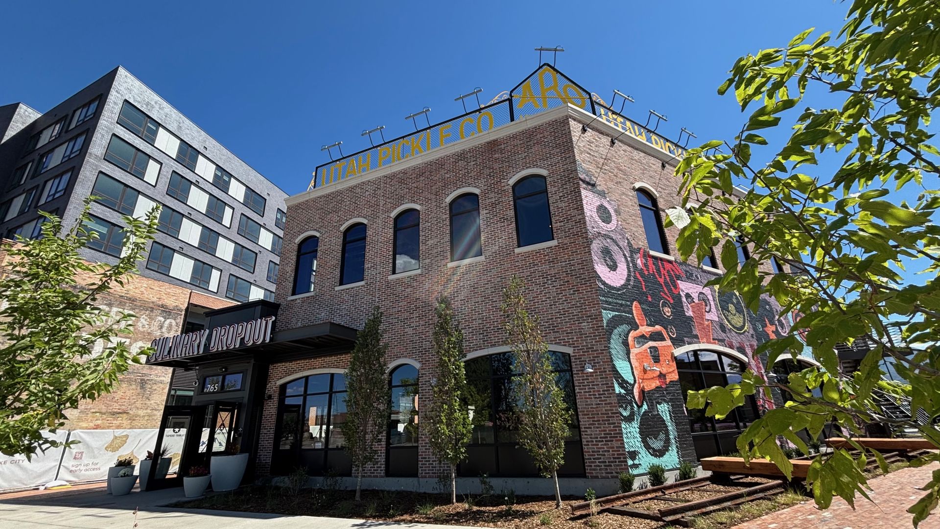 Sunny urban street with brick buildings; the right building features a colorful mural. A yellow rooftop sign reads UTAH PICKLE CO. Green trees border the scene under a bright blue sky.