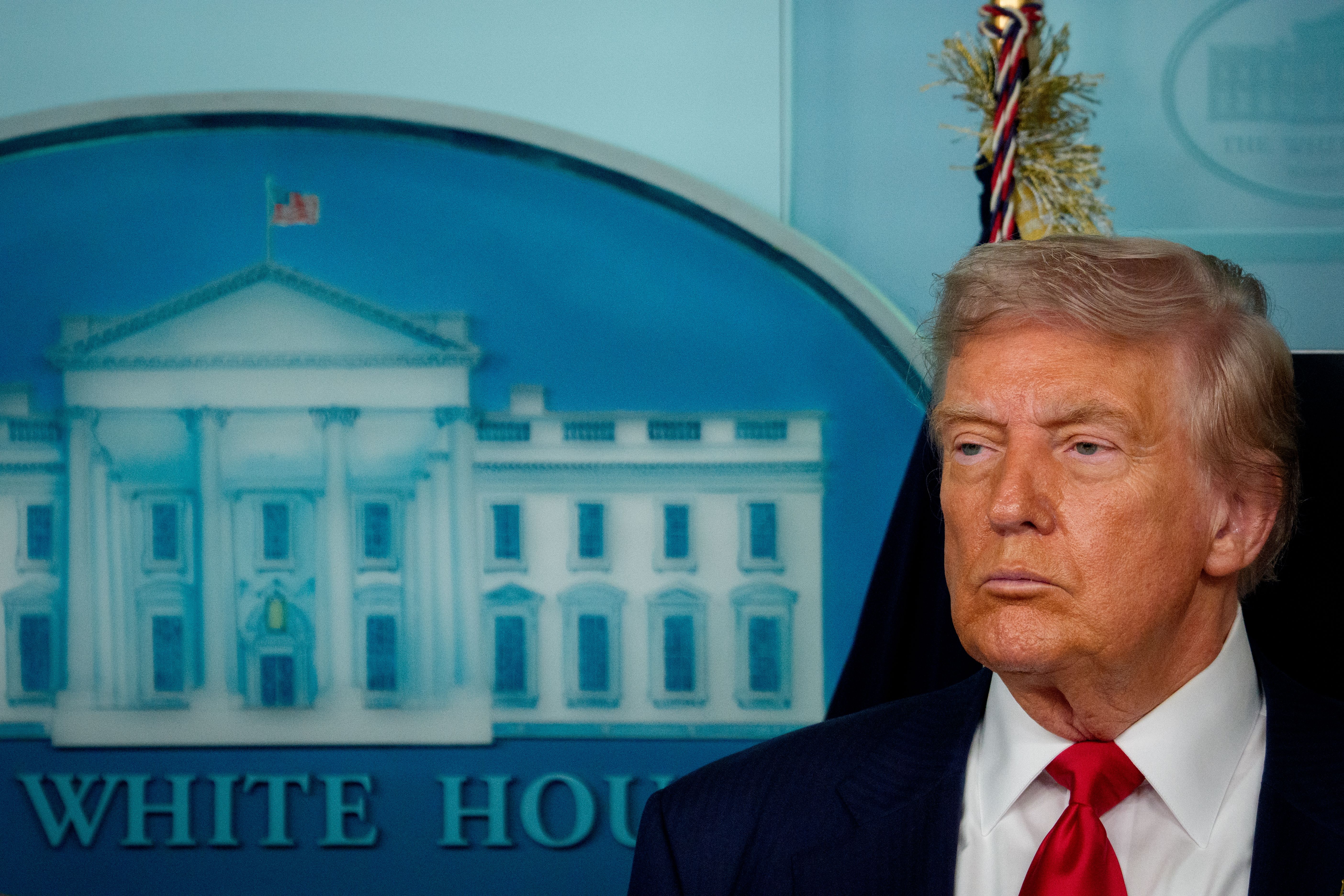 Former U.S. President Donald Trump standing with a serious expression in front of a Blue White House backdrop, wearing a dark suit, white shirt, and red tie.