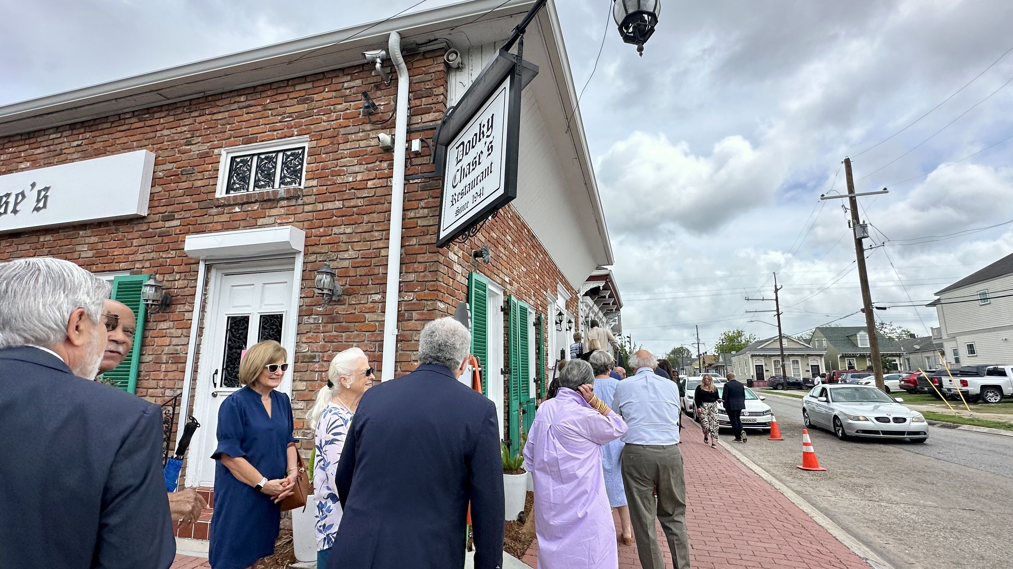 Brick building with a hanging sign reading "Dooky Chase's Restaurant" since 1941. A line of well-dressed guests stands along a red brick sidewalk with green shutters under a cloudy sky.